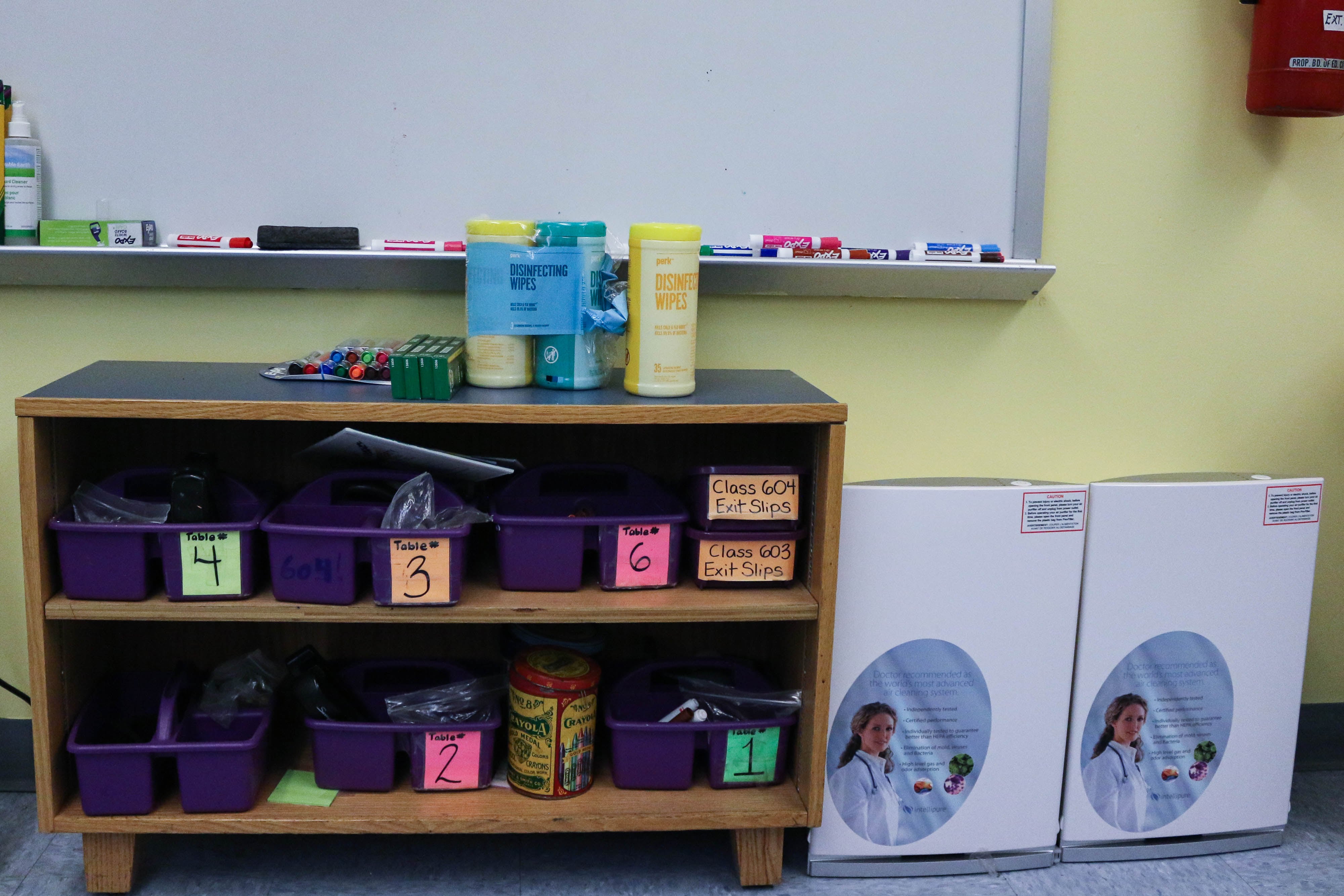 Two Intellipure air purifiers sit next to a storage shelf and a whiteboard in a New York City classroom.