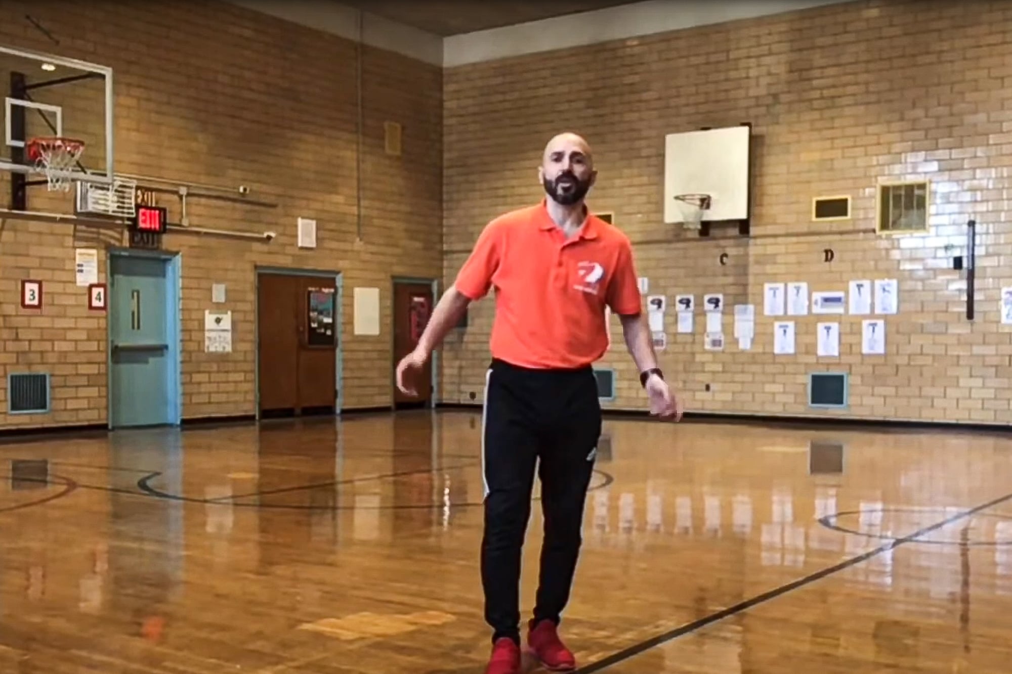 A man in a red polo shirt and black sweatpants stands in a school gym.