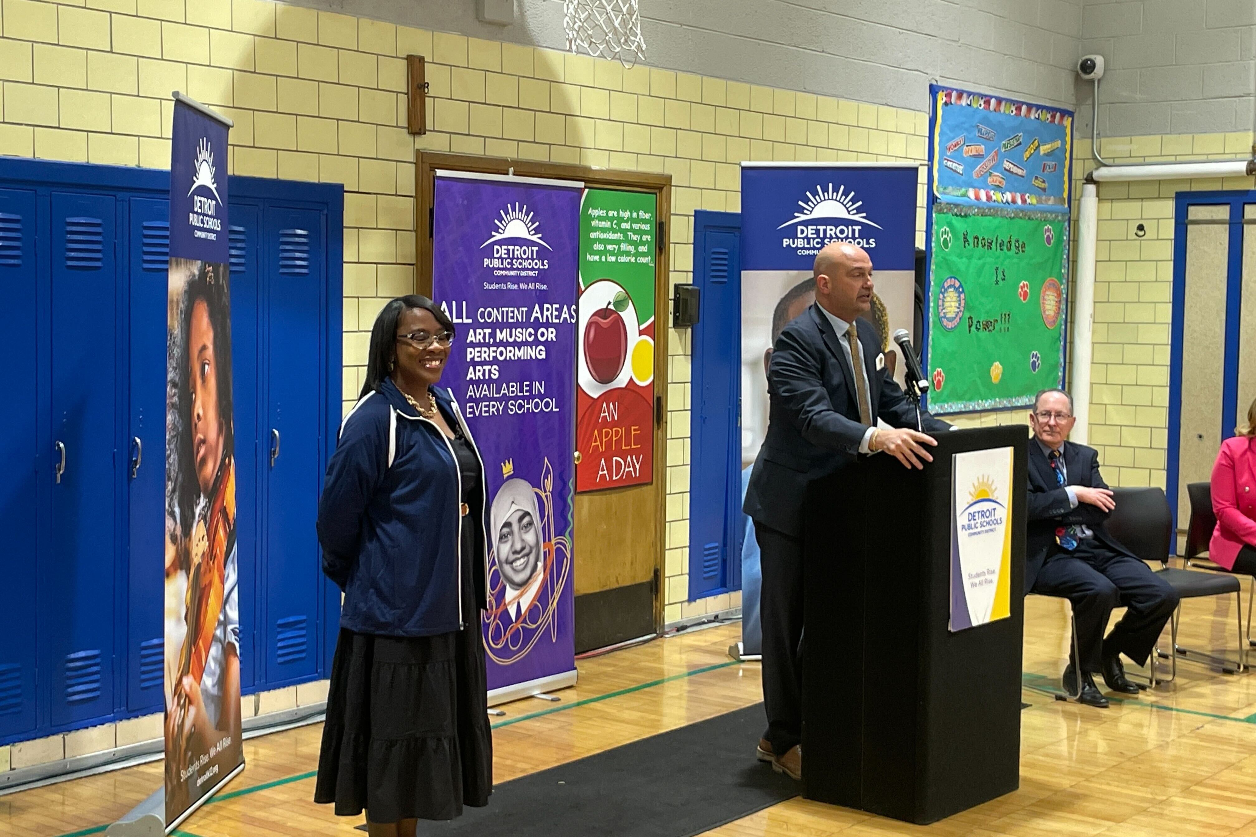 A woman wearing a black dress and a dark blue jacket stands to the left of a man in a suit standing at a lectern. Behind them are colorful flags.