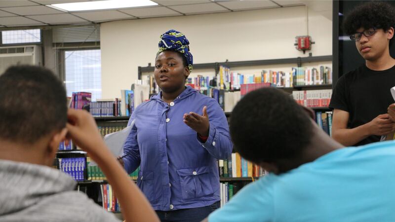 A Black girl in a blue shirt talks to a group of students in a high school. She’s in front of a bookshelf.