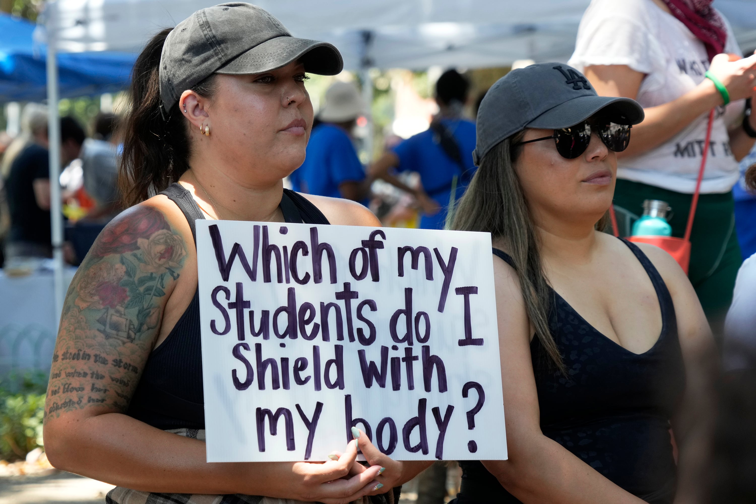 Two woman wearing baseball hats stand next to each other outside and one is holding a white sign with black words that read "Which of my students do I shield with my body?"