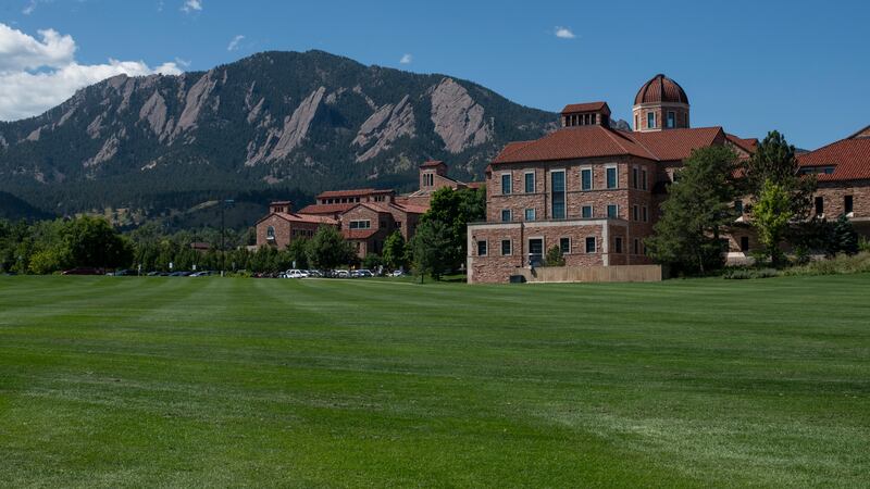 A large brick building with a red roof sits on a large lawn with green grass and the Flatirons mountain formation in the background.