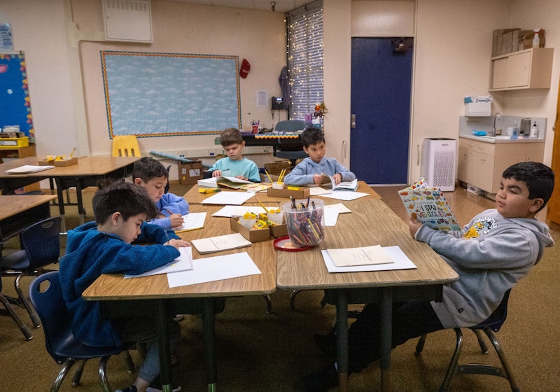 Elementary students read in a classroom. Desks are pushed together so that the students sit facing each other.