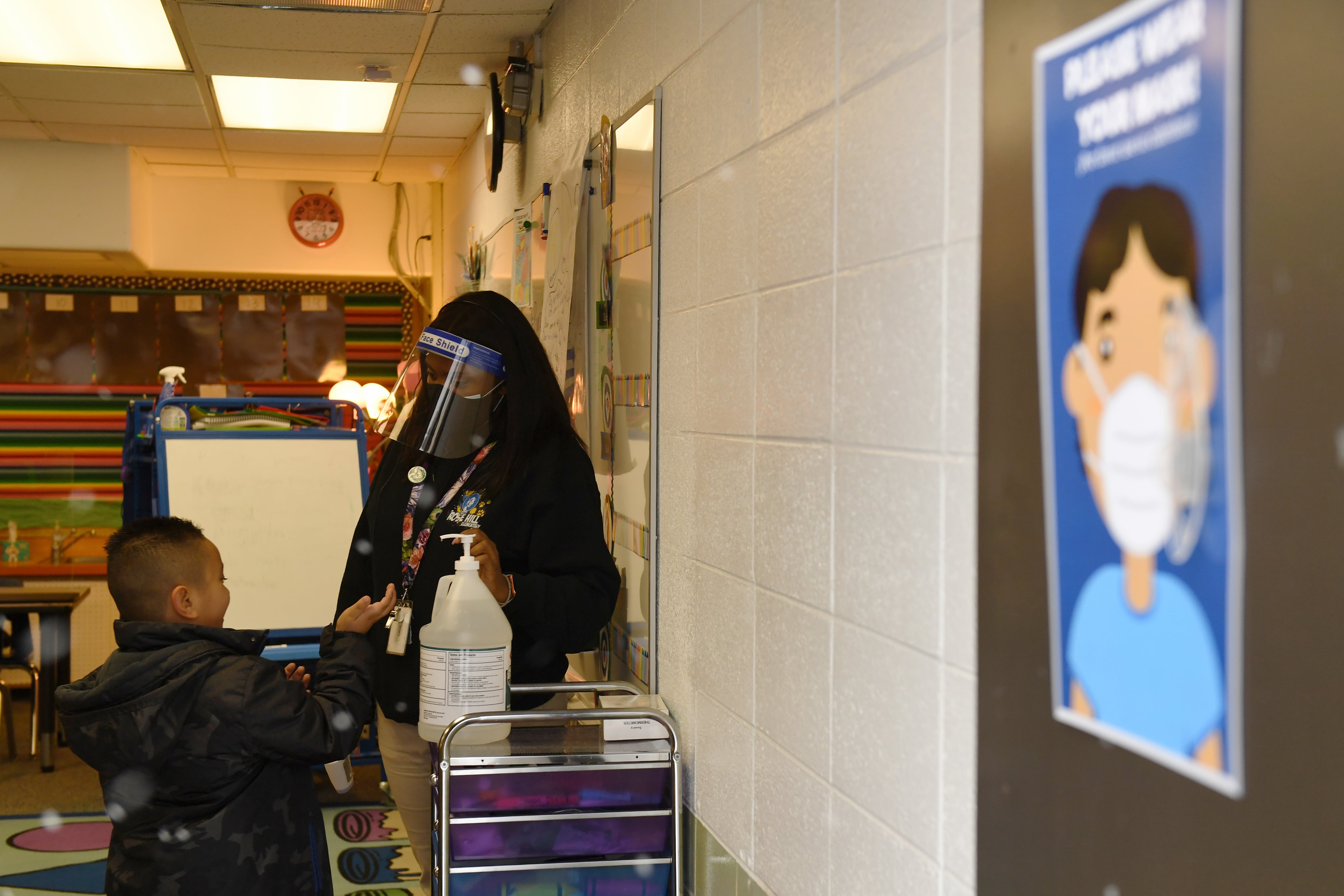 A teacher wearing a mask and face shield helps a boy sanitize his hands before class starts. A sign on the wall encourages mask wearing.