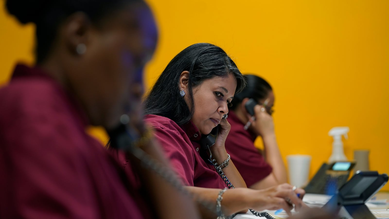 Three women wearing dark red shirts work on the phone in a call center. The woman in the center is the focal point, who appears to be typing as she is on the telephone.