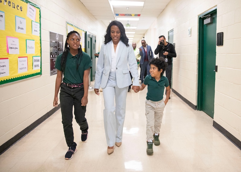 An adult wearing a blue suit walks between two young students, holding the hand of one student on the right in a hallway with white walls and green doors.