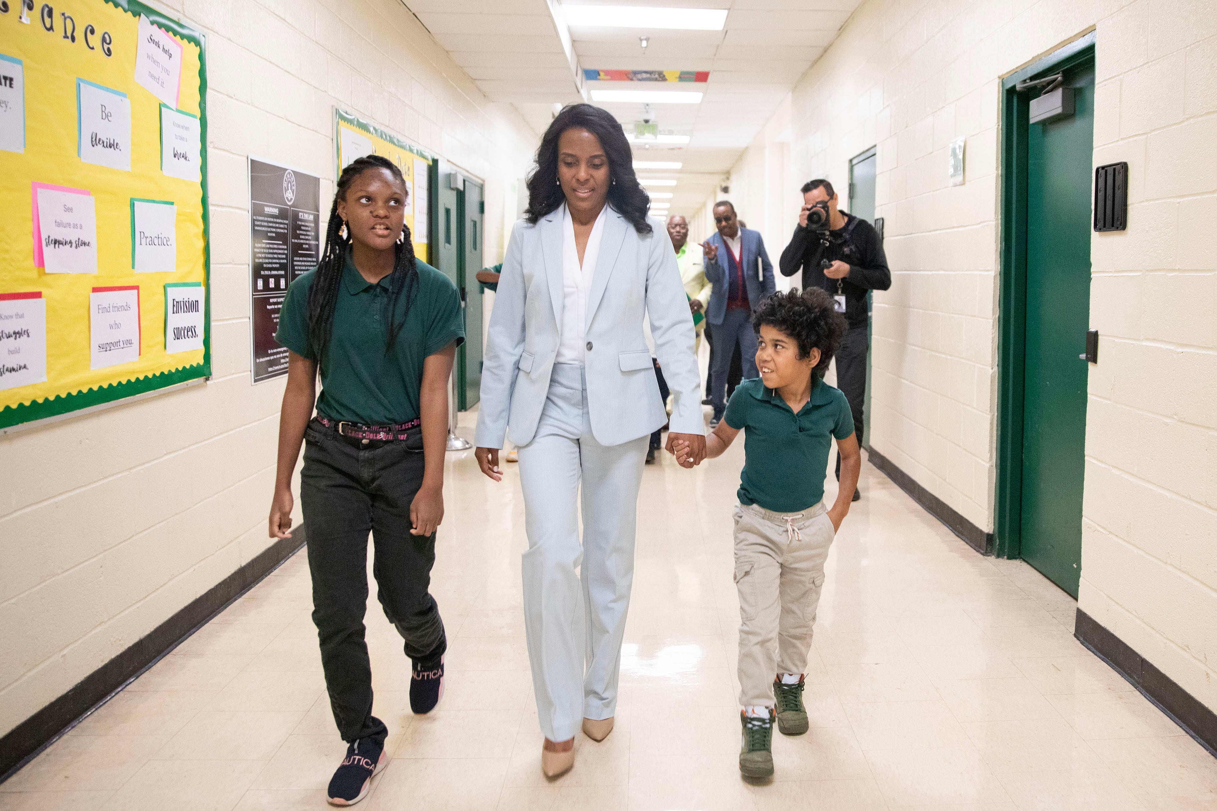 An adult wearing a blue suit walks between two young students, holding the hand of one student on the right in a hallway with white walls and green doors.