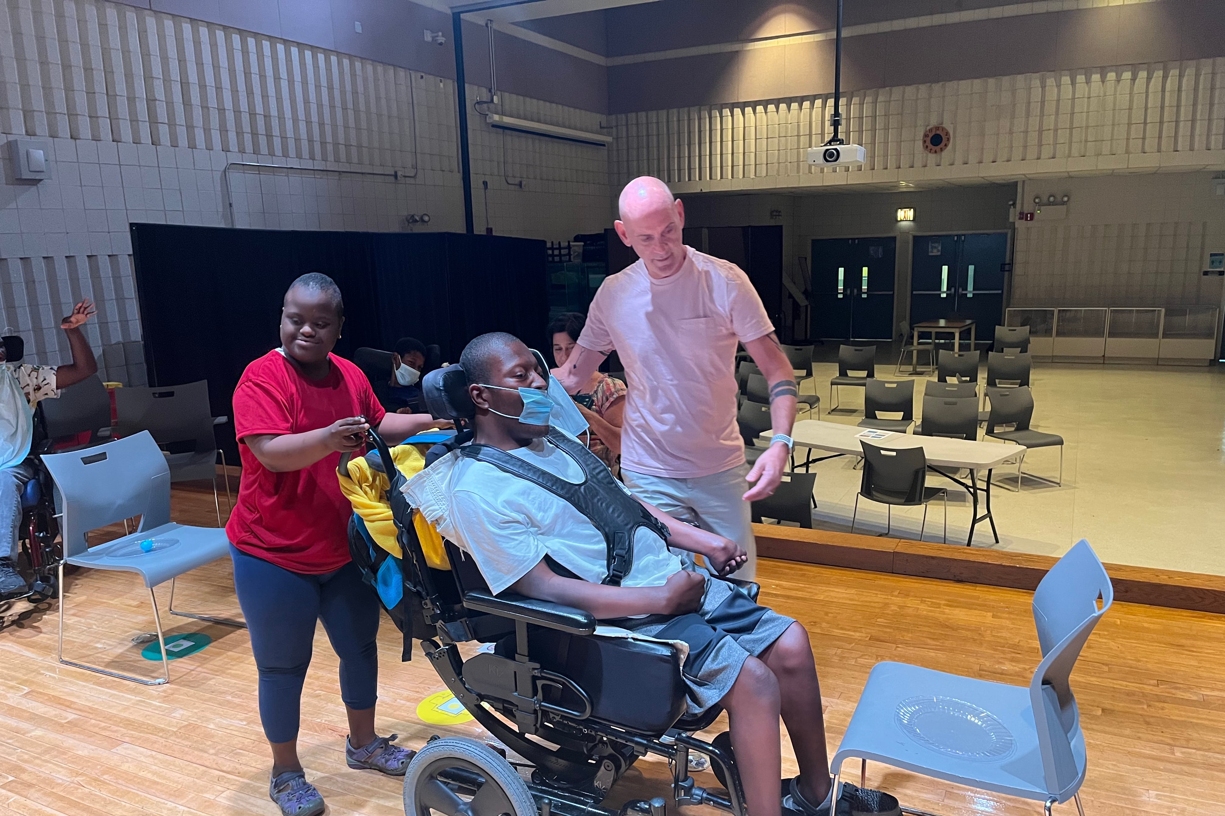 A teacher in a T-shirt and jeans stands on a stage directing two teen students, one standing in a red T-shirt and blue pants pushing the other who is in a wheelchair and wearing a white T-shirt and blue shorts.