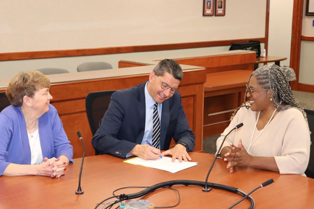 Three people in business clothes are at a wooden table in a conference room. The man in the middle is signing paperwork.