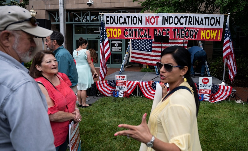 A group of people standing on a green lawn outside in front of a homemade sign that is critical of "critical race theory" or CRT.