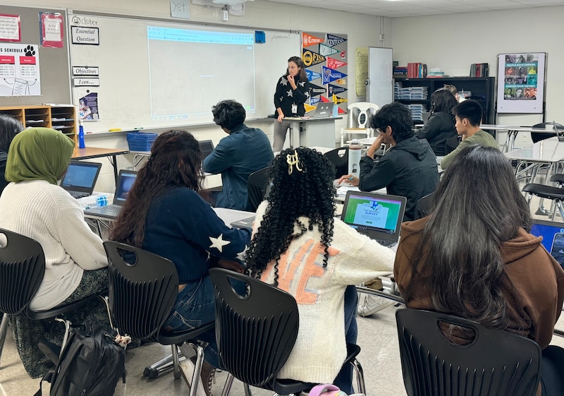 A teacher stands in the background next to a dry erase board with a group of students sitting in their chairs in the foreground.