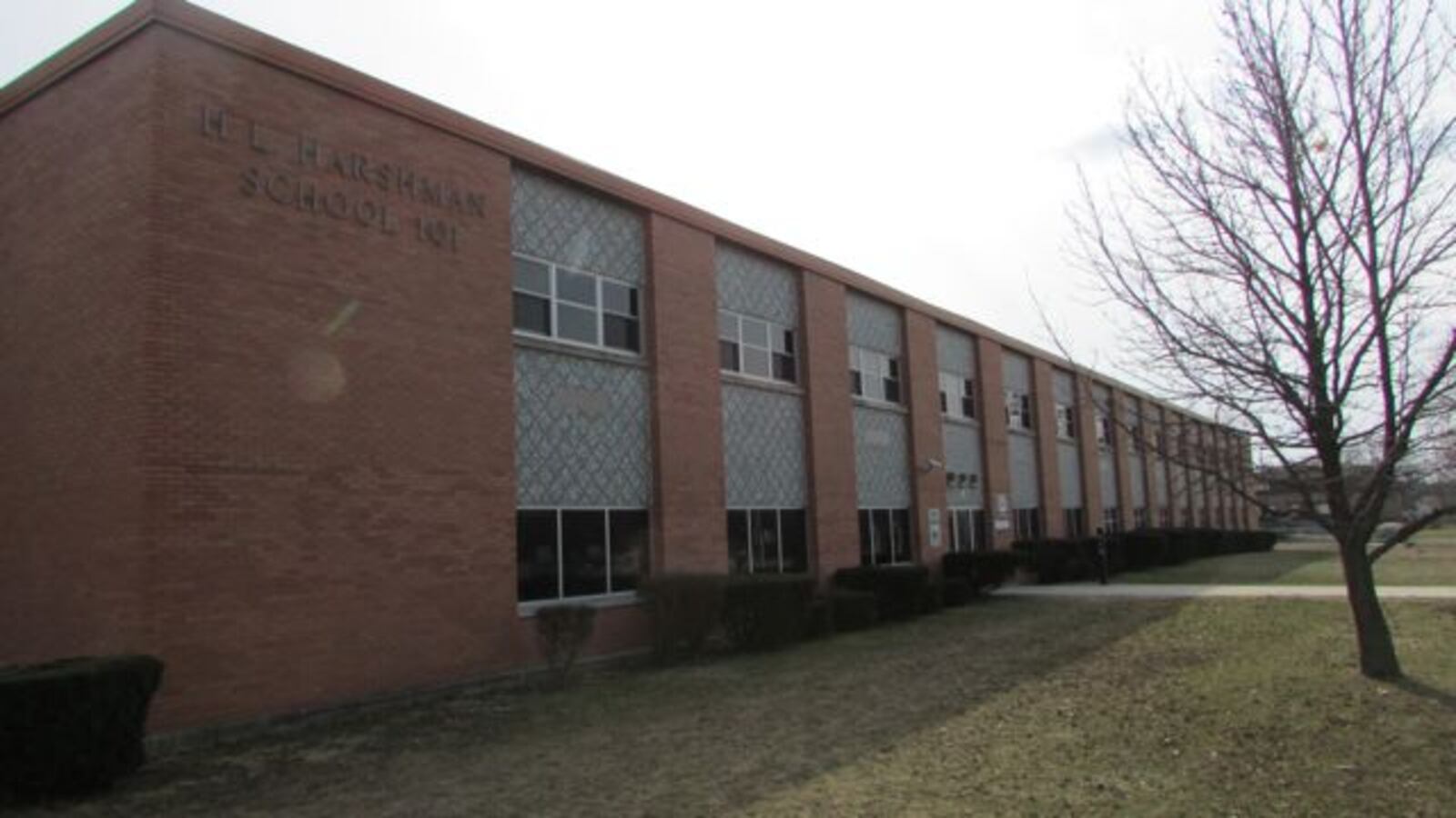 A brick building with a sign that says “H.L. Harshman School 107” sits on a green lawn with a bare tree in the foreground.
