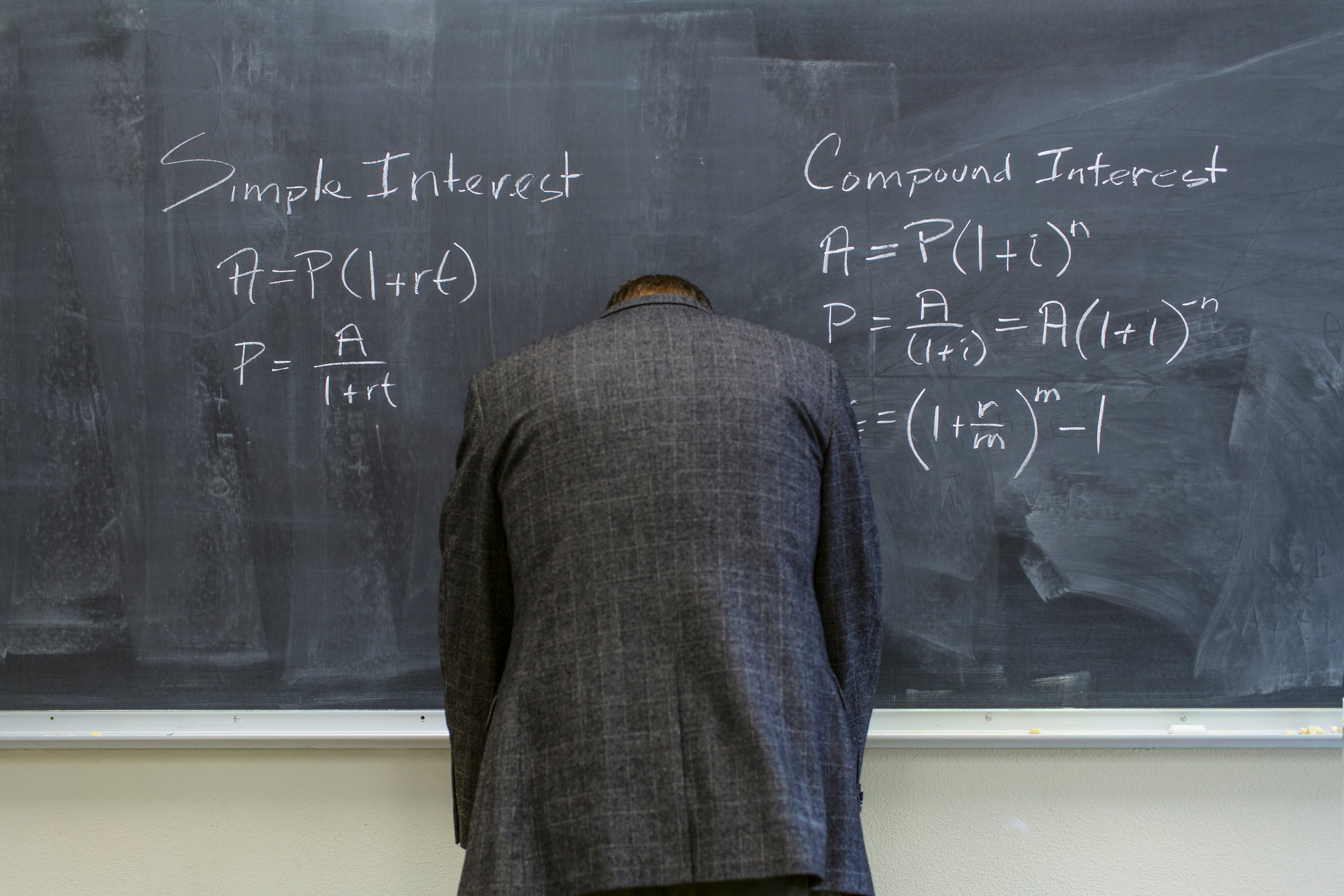 A man in a suit jacket rests his head on a chalkboard.
