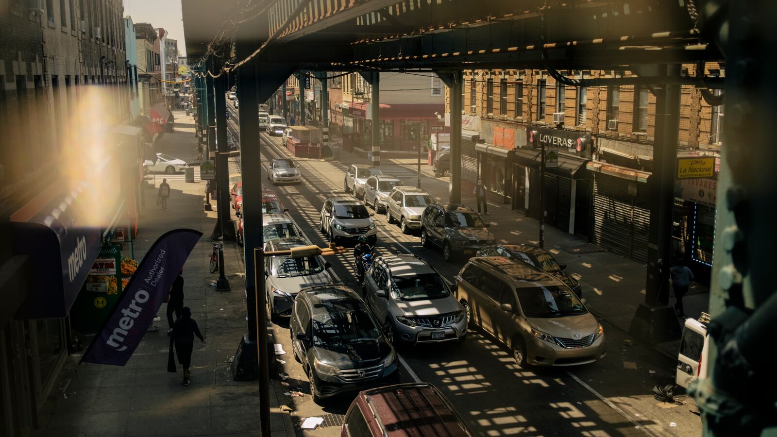 A bustling street in Cypress Hills, Brooklyn, with traffic moving under a subway track and shadows lining the street.