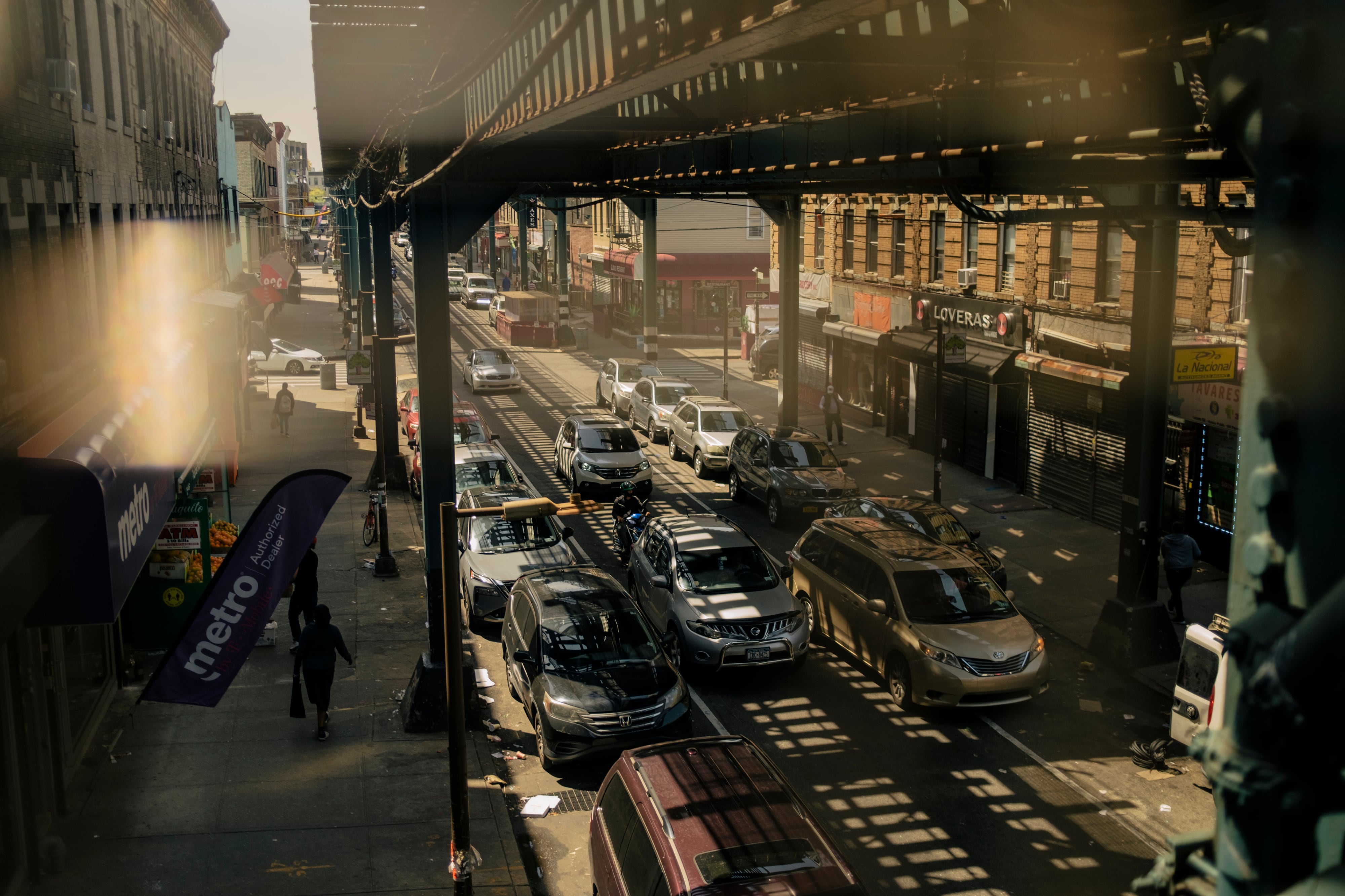 A bustling street in Cypress Hills, Brooklyn, with traffic moving under a subway track and shadows lining the street.