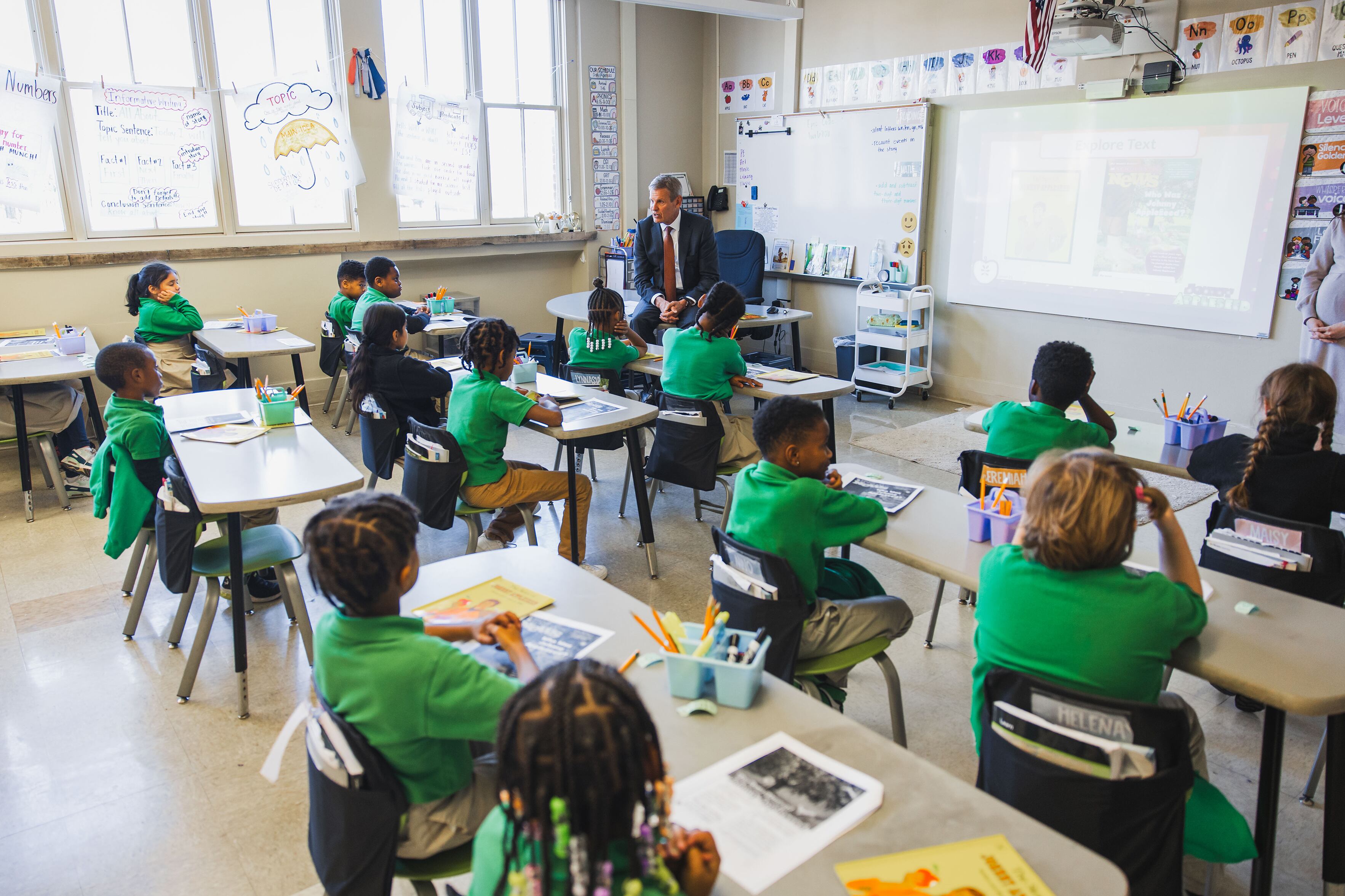 A man in a suit sits on a desk in front of a classroom with young students sit at their desks.