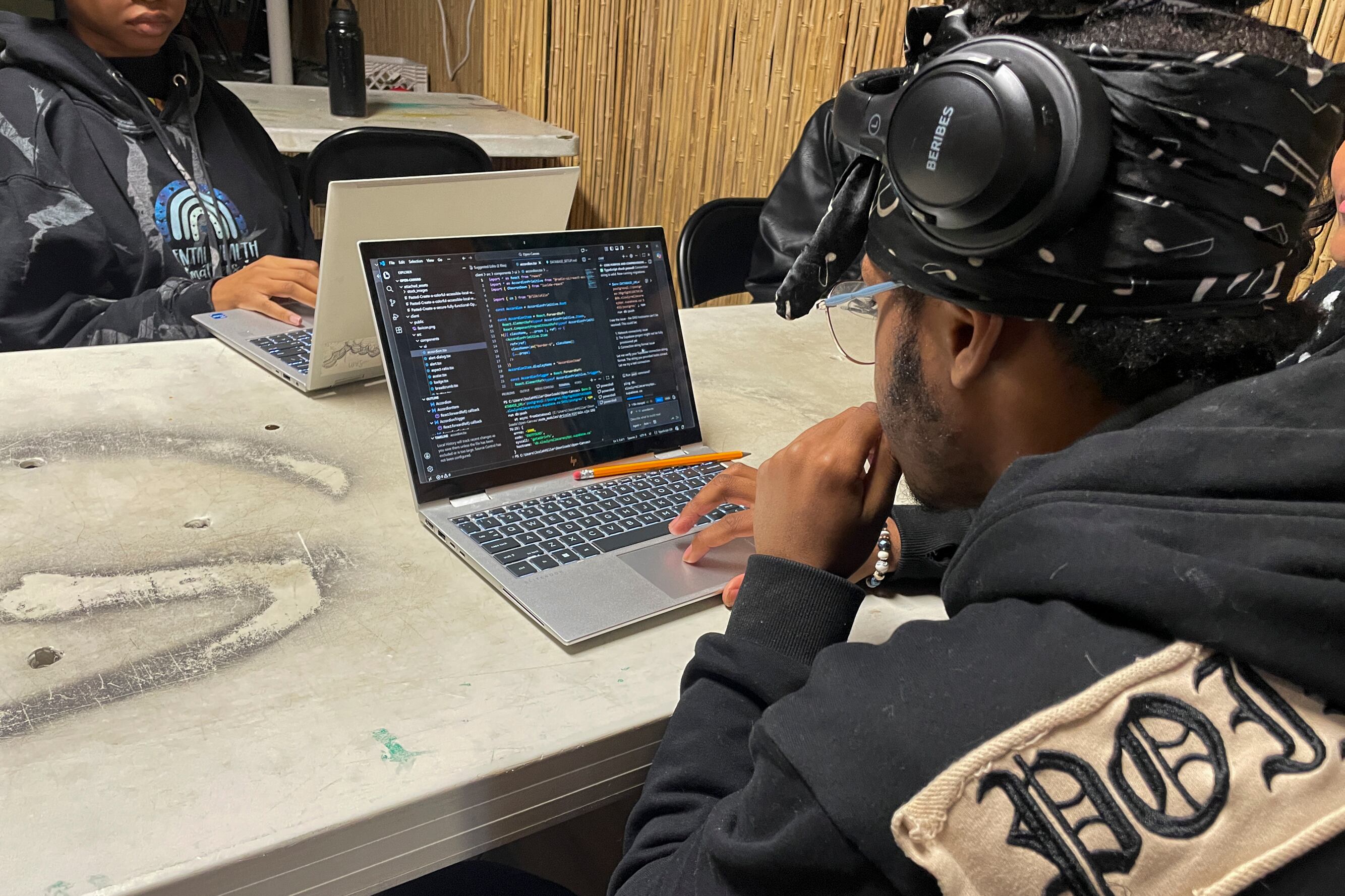 A photograph of the back of a black male looking at the laptop in front of him on the table. There are lines of code on the laptop's screen.