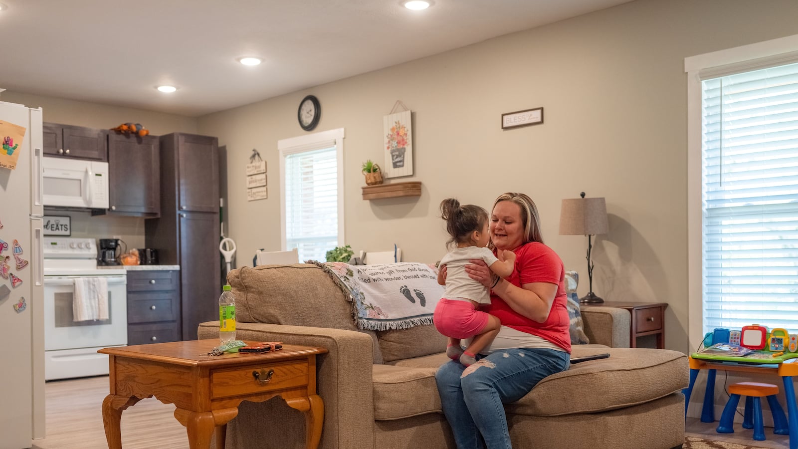 A woman holds her toddler in her arms as she sits on the couch of their apartment.