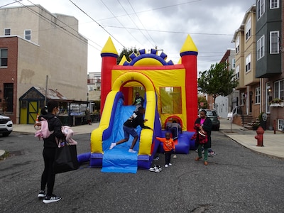 Children play on a bouncy castle that is in the middle of a street while an adult watches out for them. Buildings and a cloudy sky are in the background.