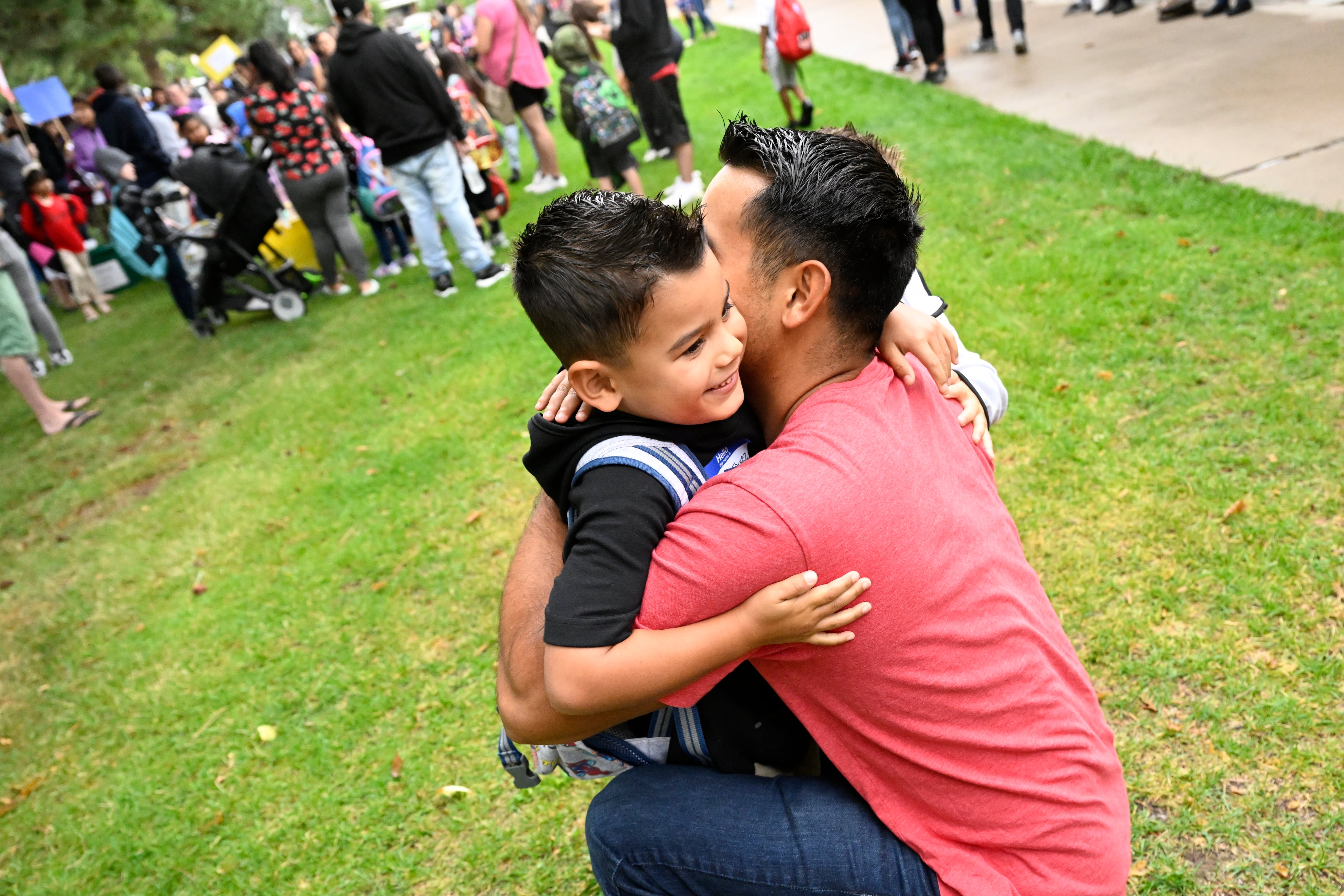 A young smiling boy in a black shirt hugs his father. The father is wearing a red shirt and crouches down to be at his son’s level. They both have gelled black hair in the same style.