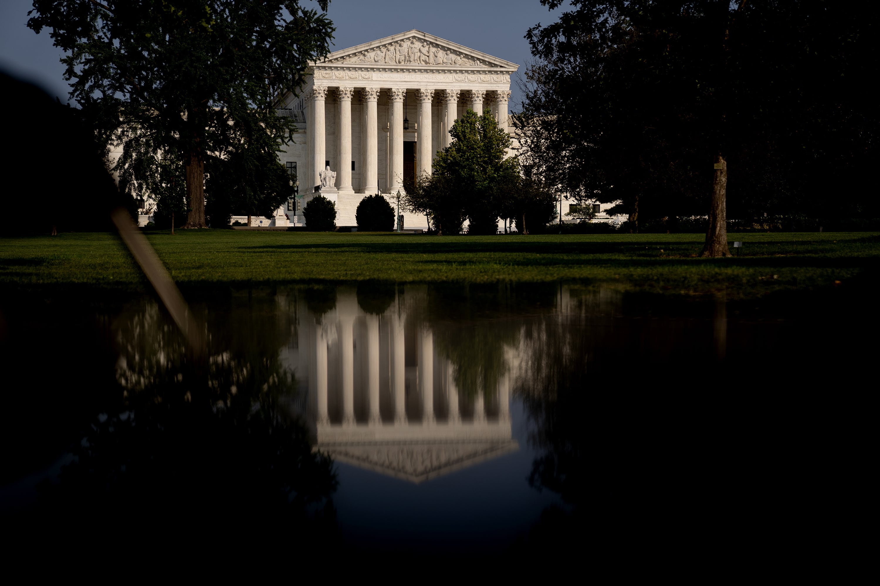 A large white stone building is seen through green trees and a reflection of the building on a pond.