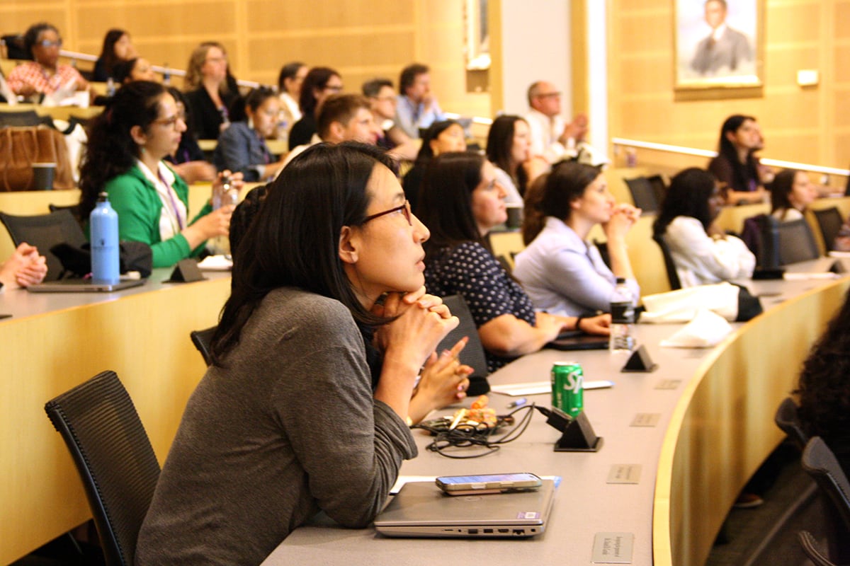 A woman in a gray top and glasses is in the foreground of a crowd of people listening to a speaker.