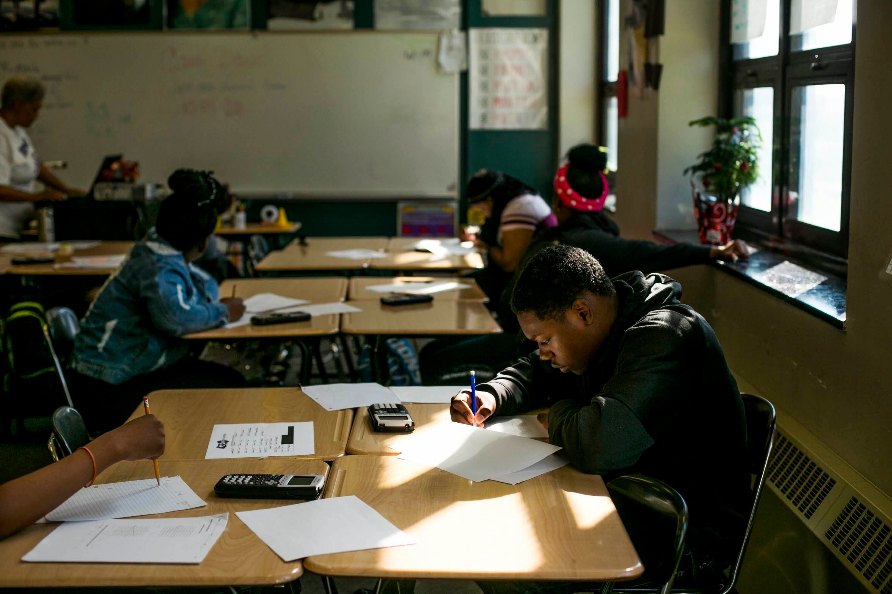 Several high school students use pencils and calculators to complete assignments in a classroom. Light streams in from windows on the right of the photo.