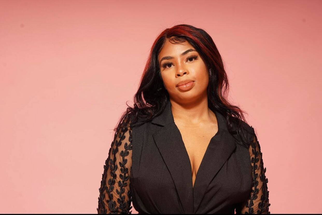 A Black woman wearing a black blouse looks into the camera as she stands in front of a pink wall.