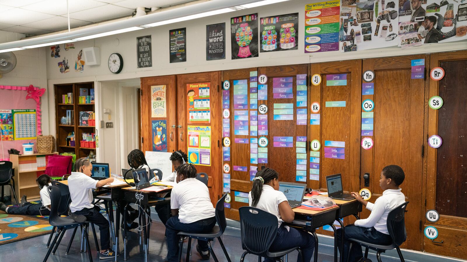 Two groups of students sit at tables and work on laptops.