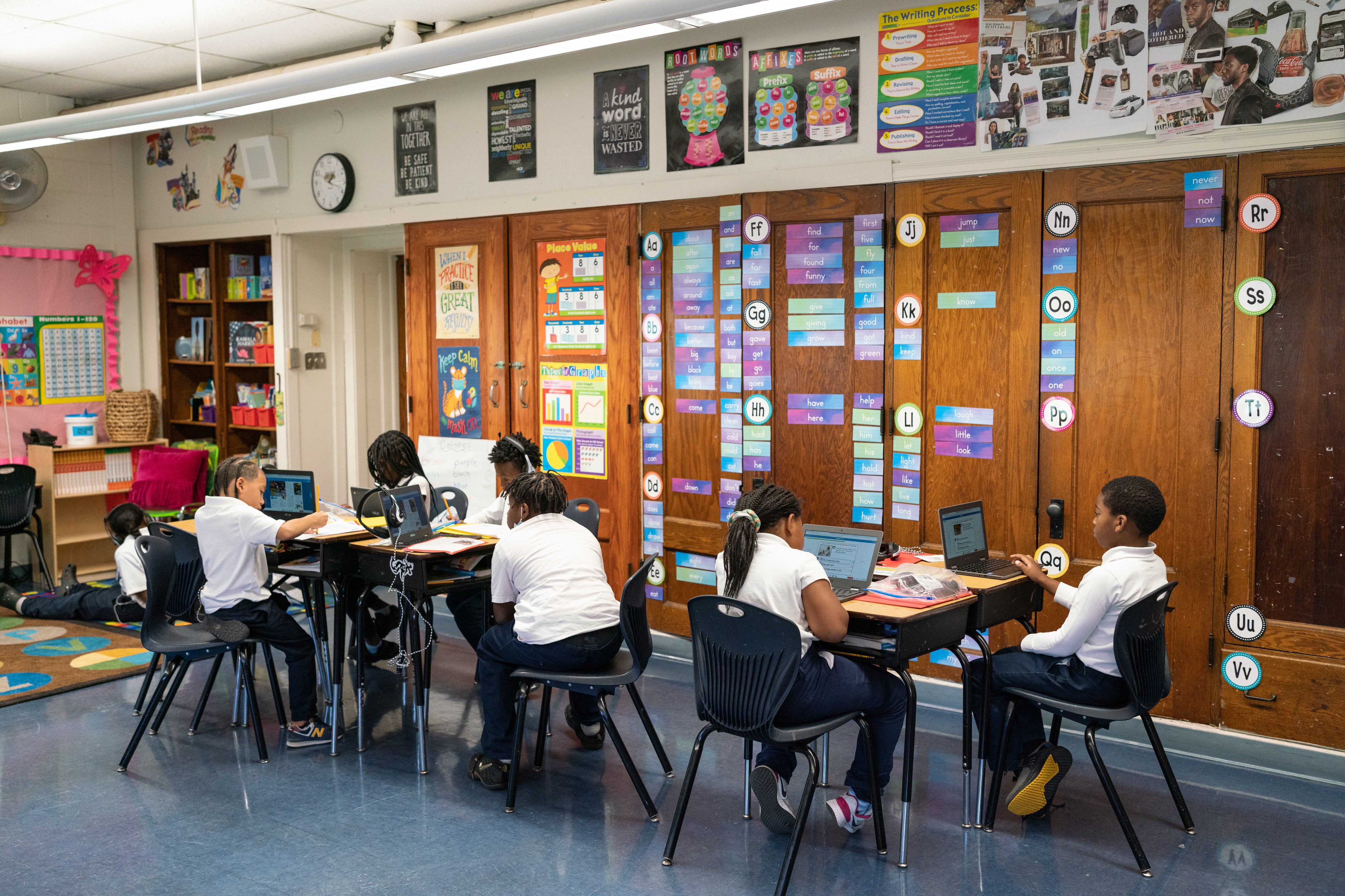 Two groups of students sit at tables and work on laptops.