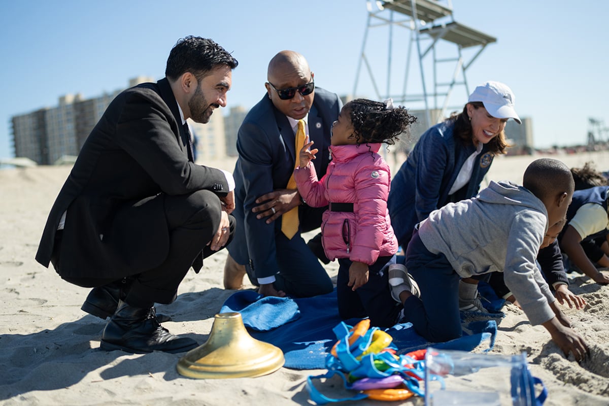 Two men in dark suits talk to a little girl in a pink jacket on the beach.