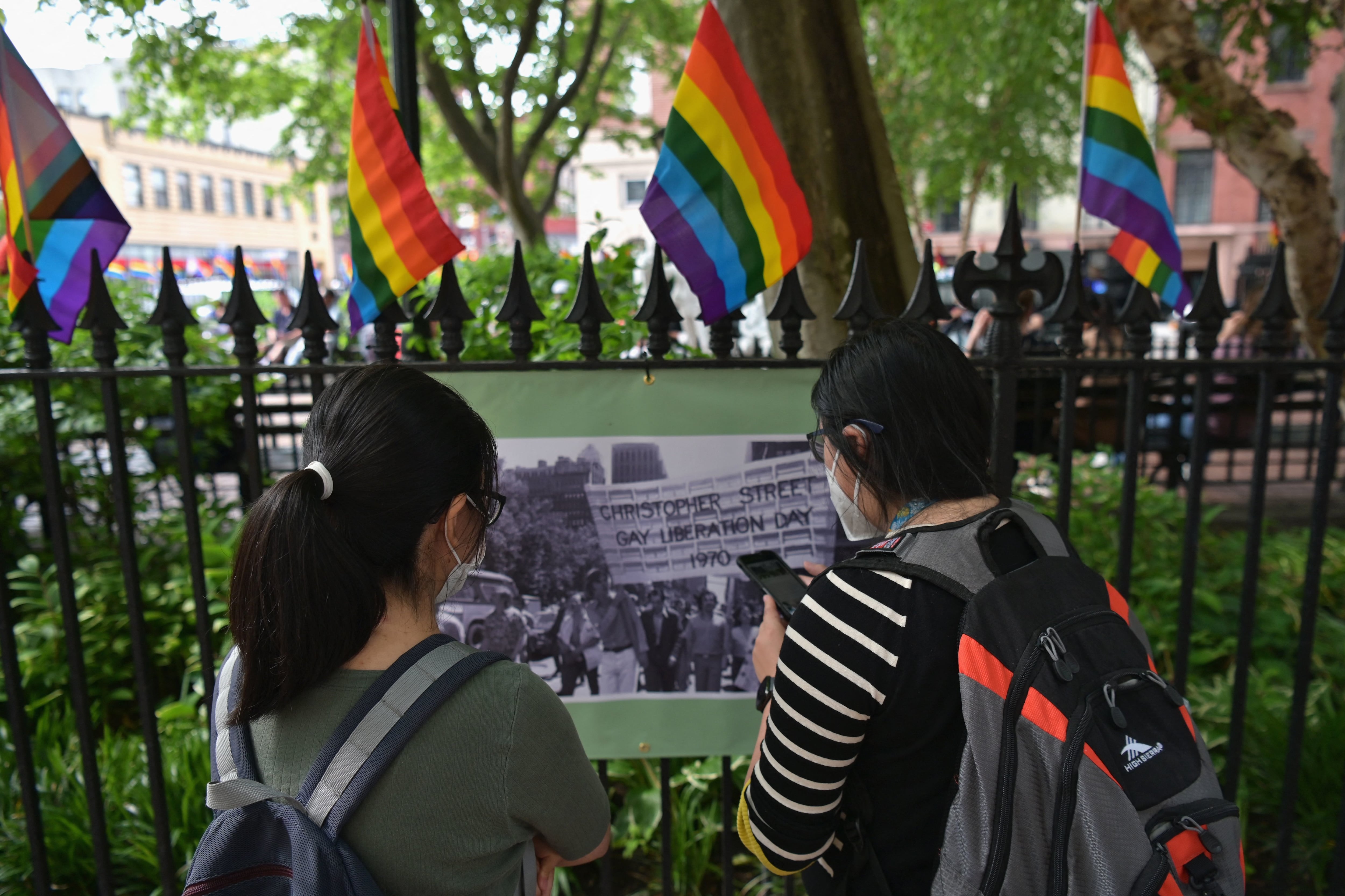 Two young women look at a historical photograph outside of the Stonewall National Monument in New York City, with Pride flags marking the top of a metal fence.