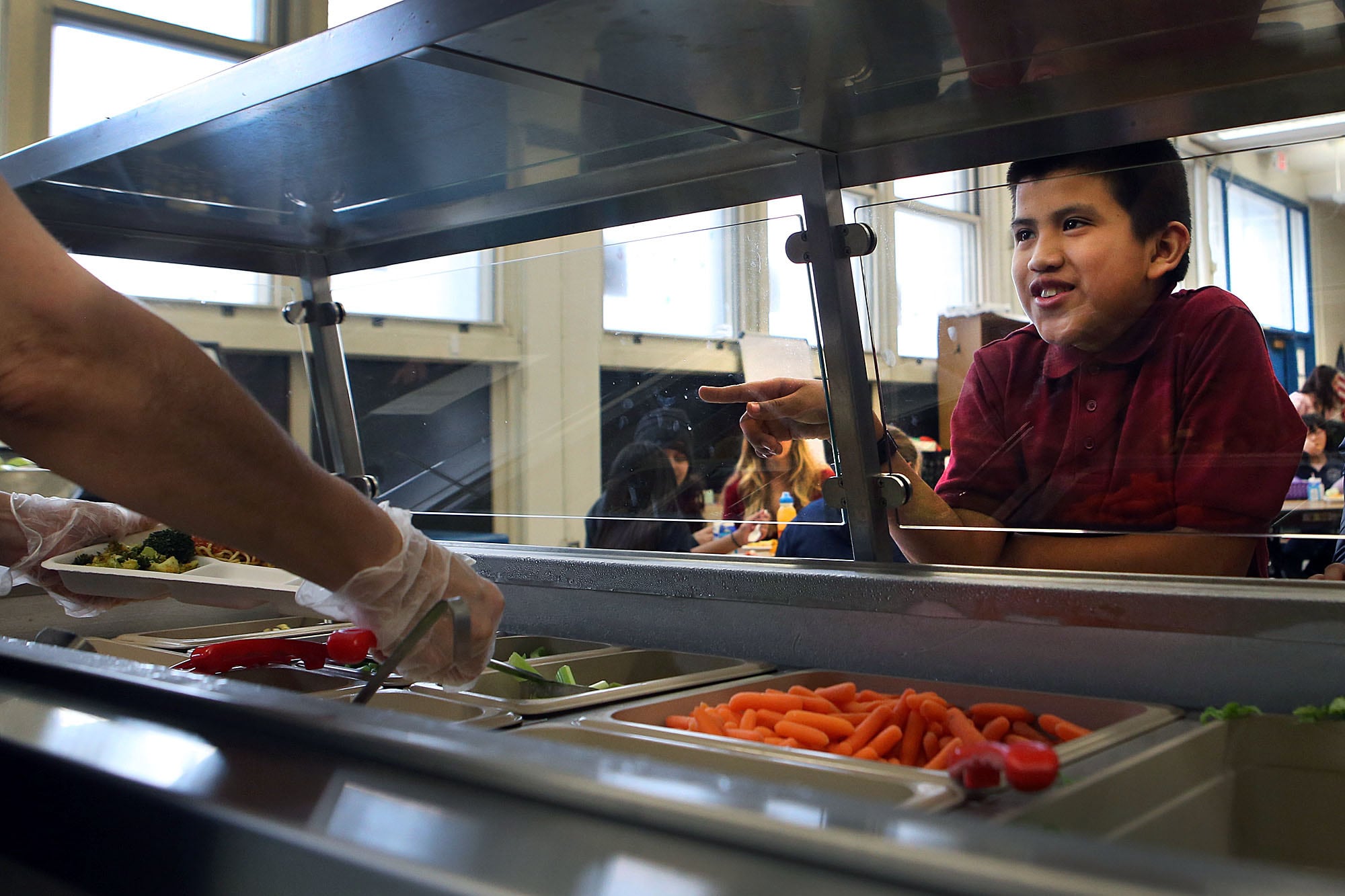 A young student wearing a red shirt points and smiles through a glass sneeze shield to food while an adult serves food on a tray.