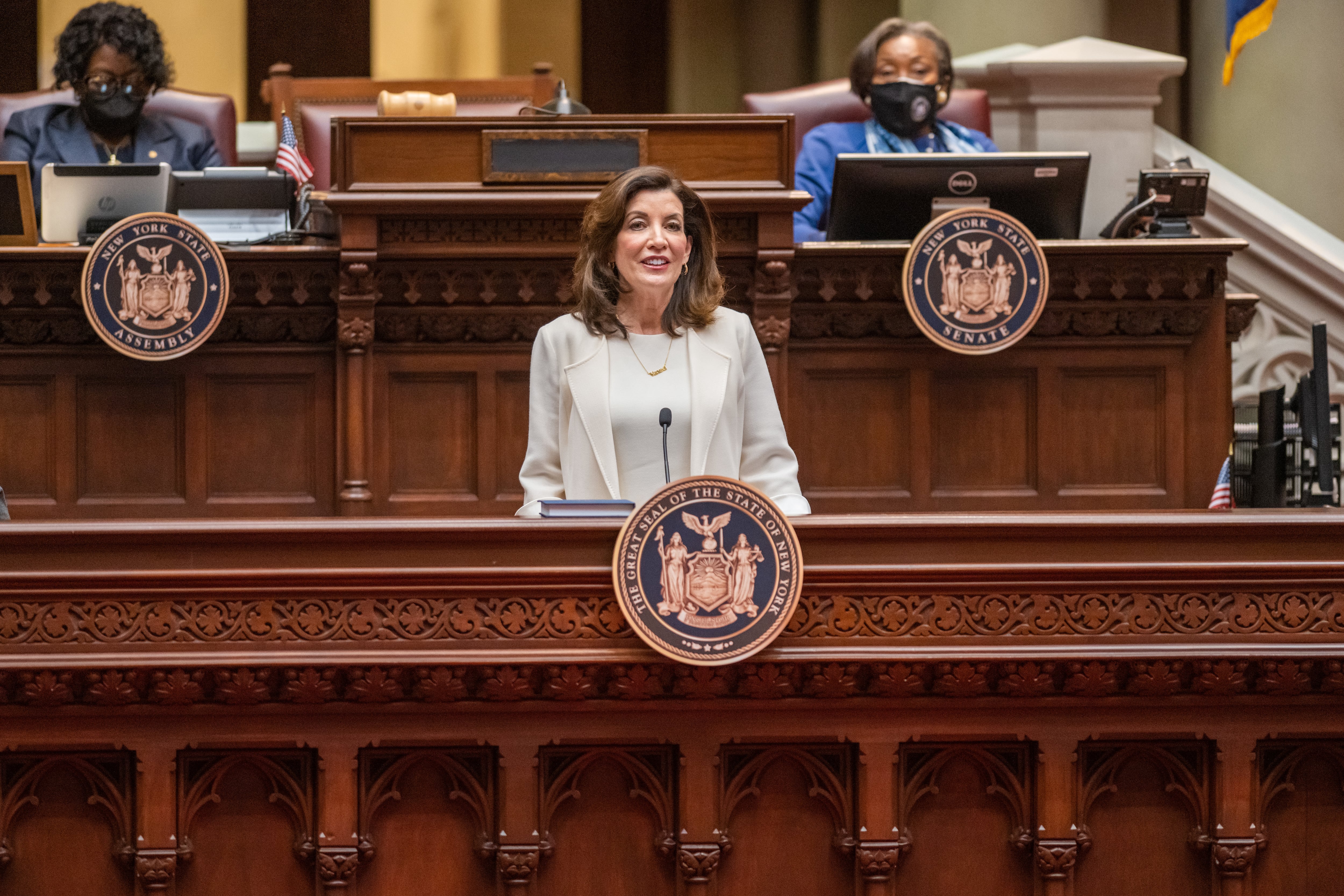 A woman stands at a lectern with two other people seated in the background.