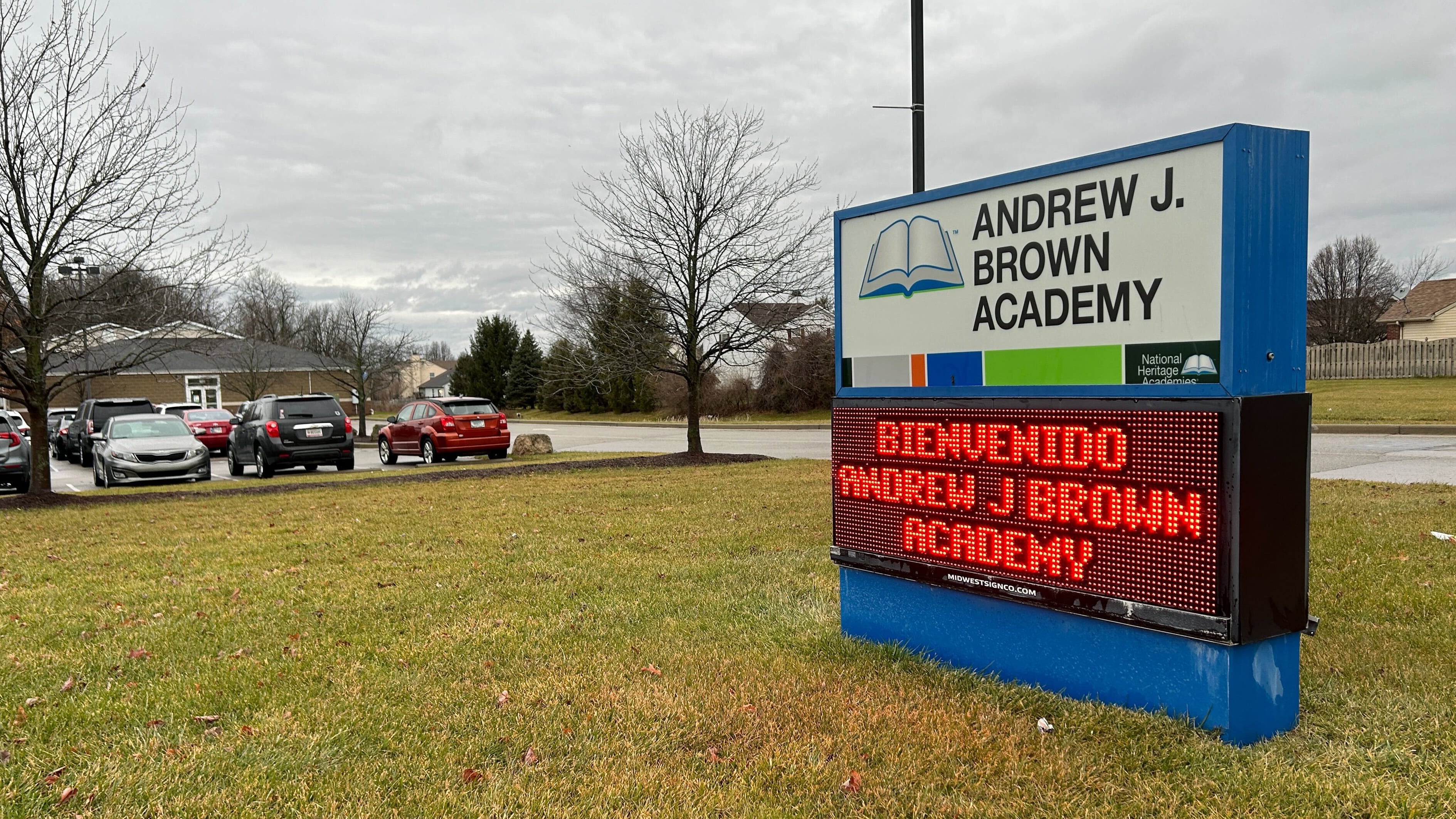 A sign that reads "Andrew J. Brown Academy" and "Bienvenidos Andrew J. Brown Academy," sits on a green lawn with cars and grey sky in the background.
