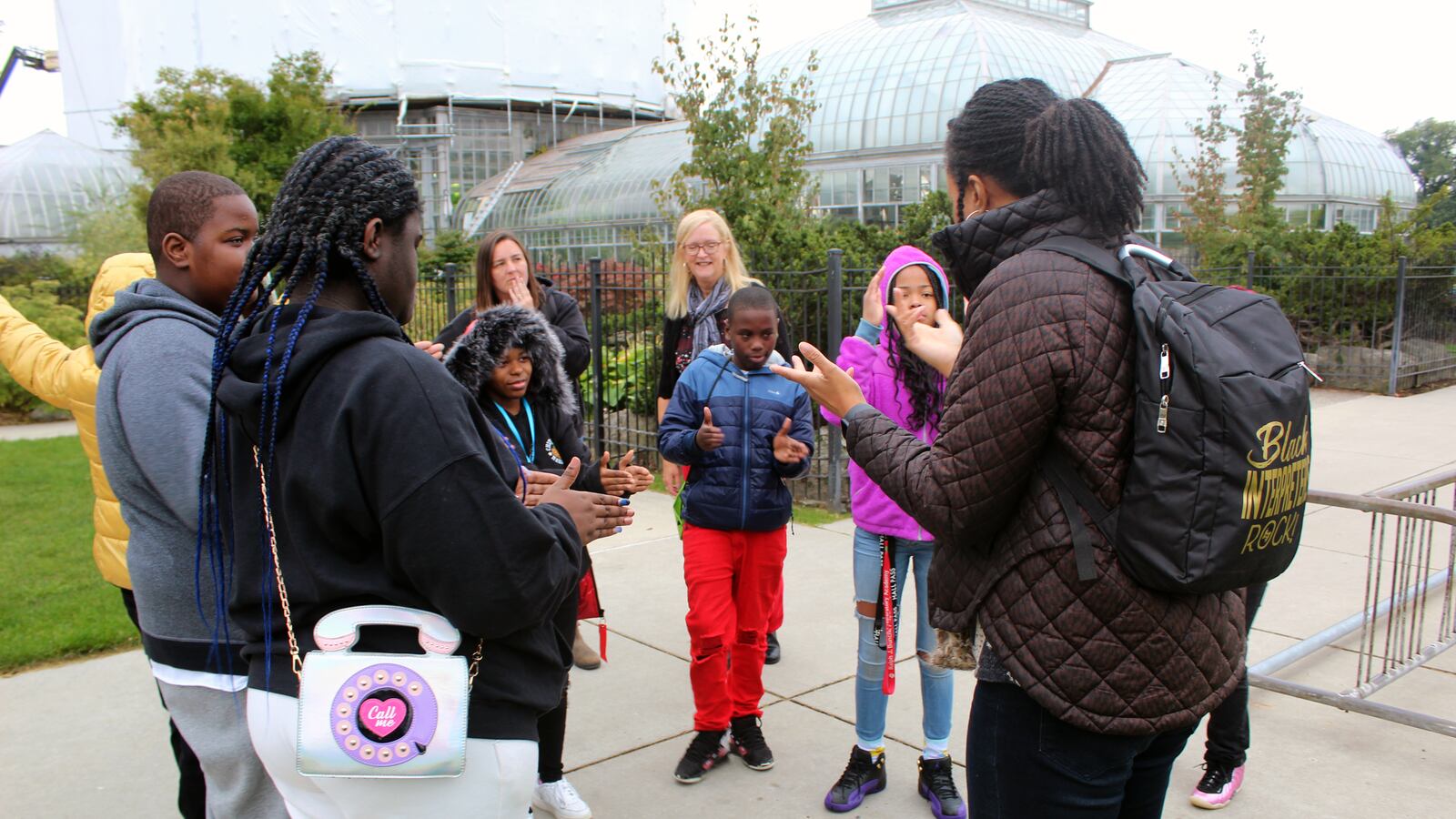 A group of students and adults standing in a circle on pavement, in front of a garden outside a glass building.