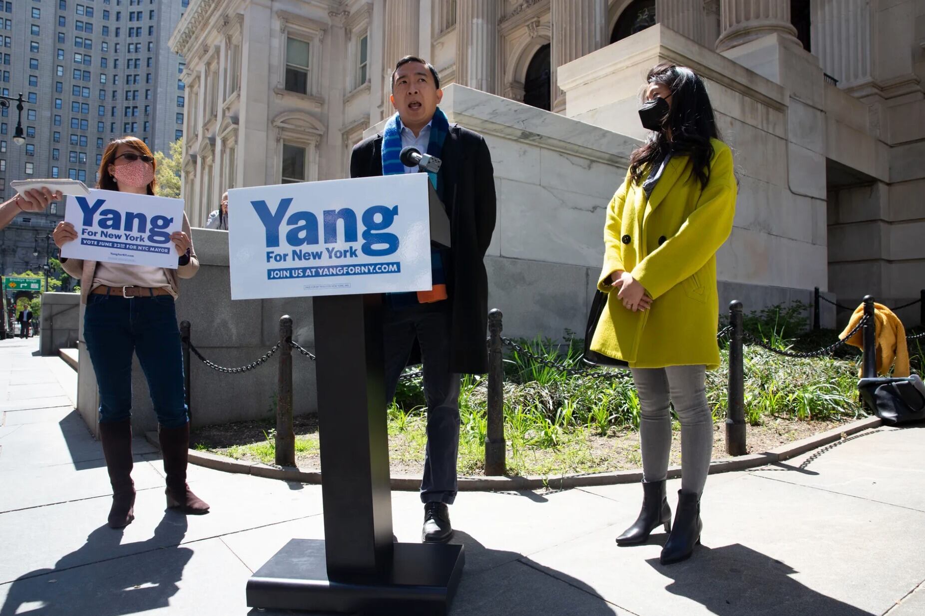 NYC mayoral candidate Andrew Yang speaks at a podium that bears his campaign sign, with a woman holding his campaign sign on his left and a woman wearing a yellow coat to his right.