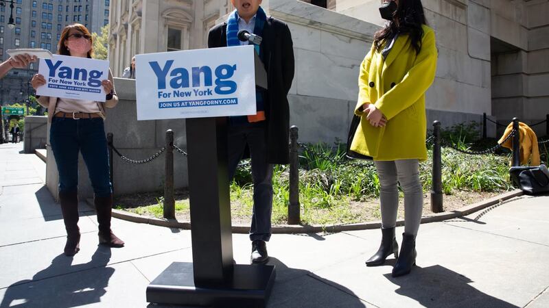 NYC mayoral candidate Andrew Yang speaks at a podium that bears his campaign sign, with a woman holding his campaign sign on his left and a woman wearing a yellow coat to his right.