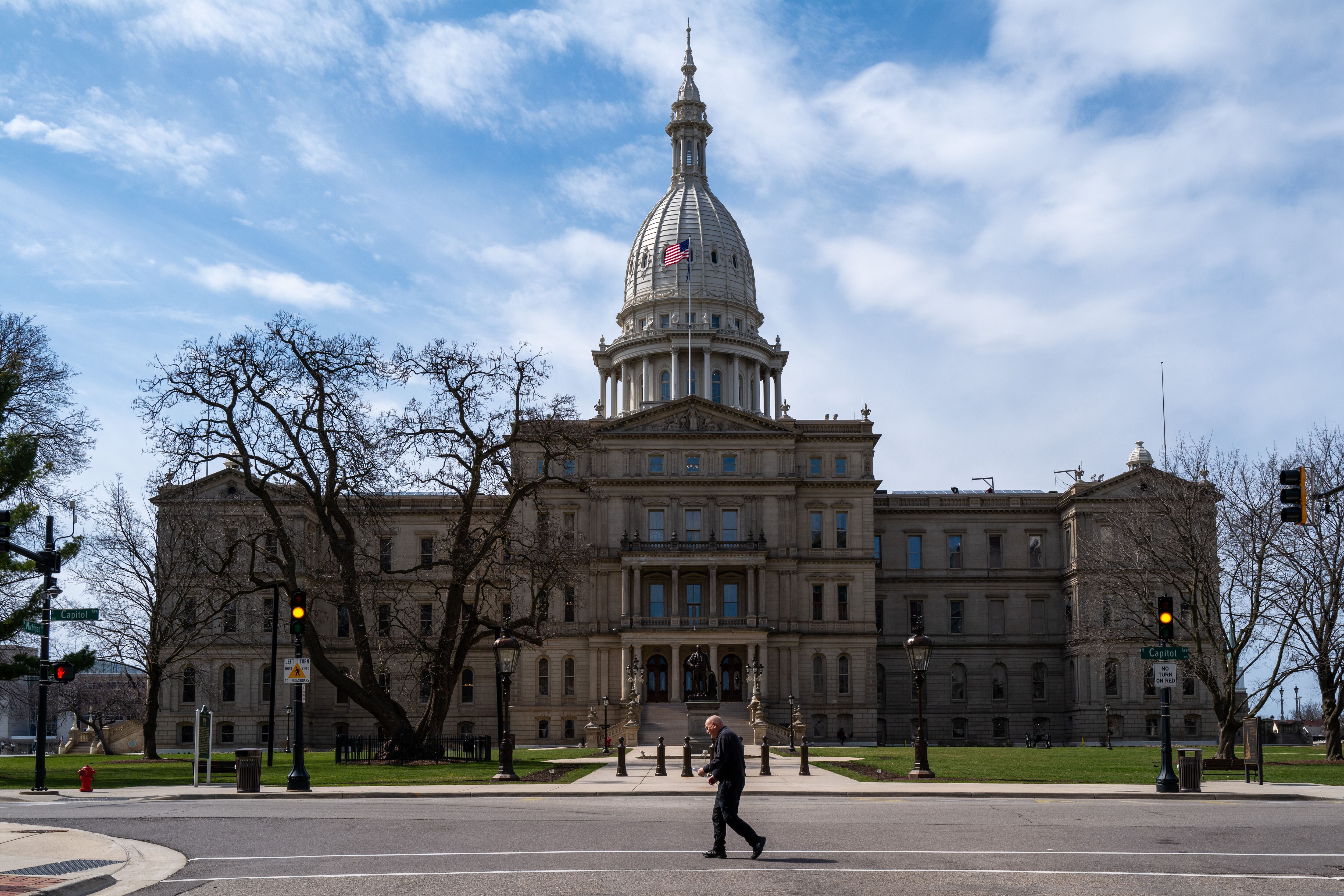 A person crosses the street in front of the Michigan Capitol with a blue sky in the background.