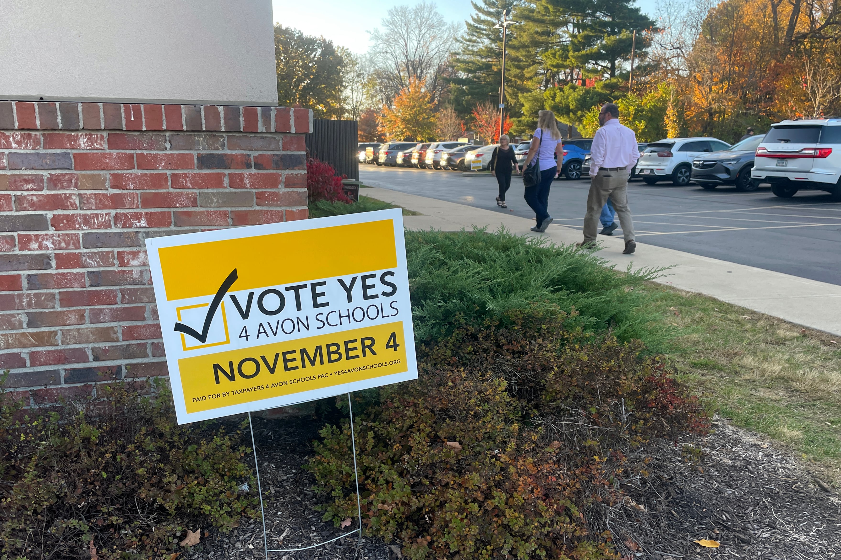 A photograph of a yellow political sign outside of a building with a group of people walking by in the background.