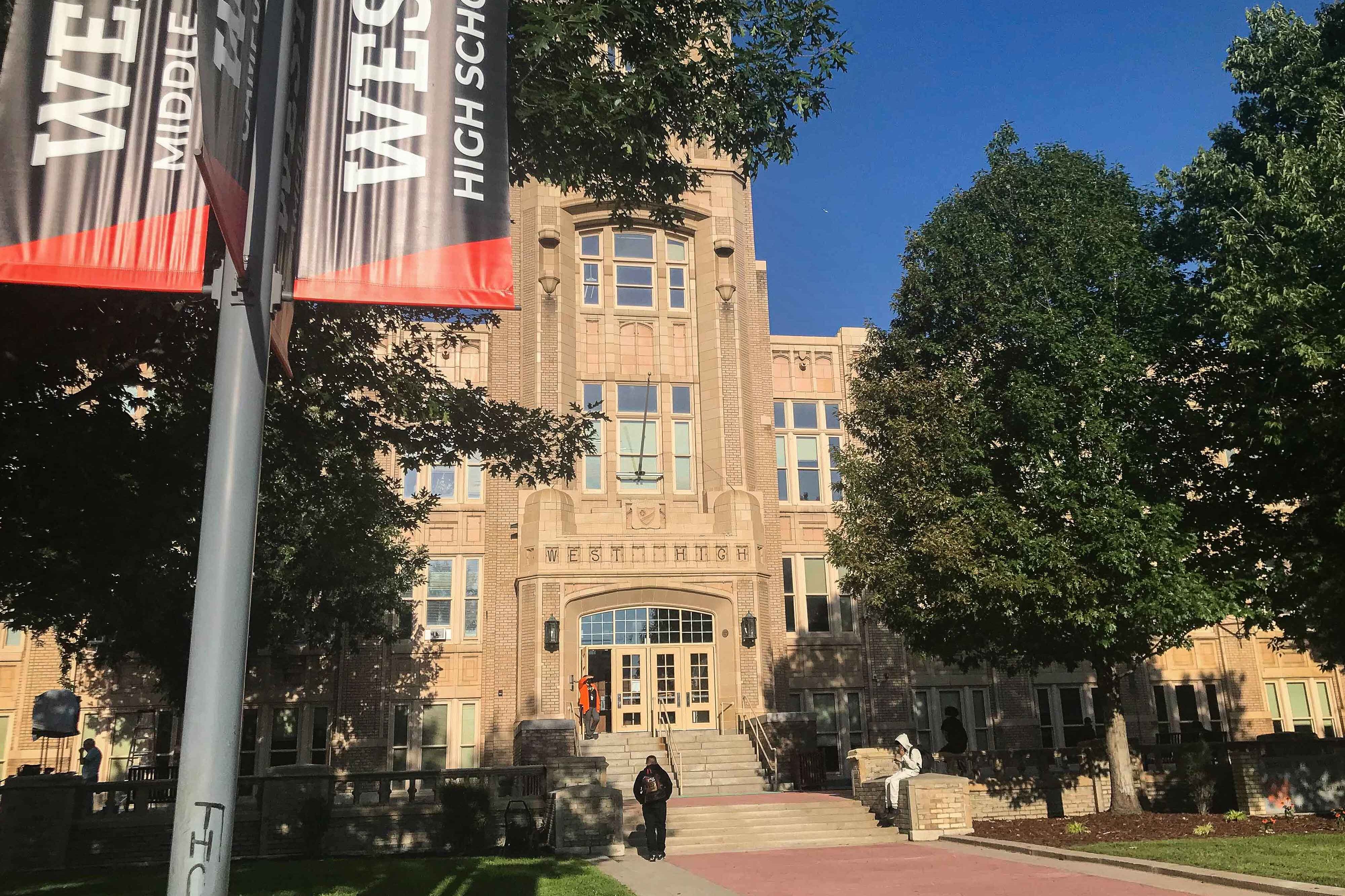 A student sits on the steps of Denver’s West High School. A black-and-orange banner in the foreground says “West High School.”