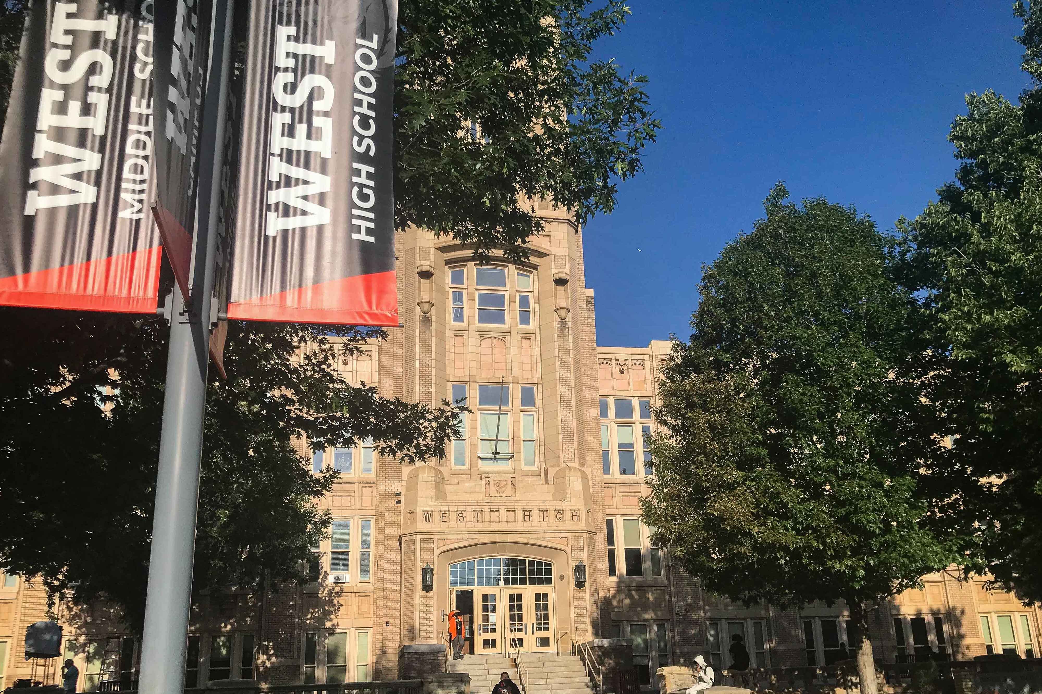 A student sits on the steps of Denver’s West High School. A black-and-orange banner in the foreground says “West High School.”