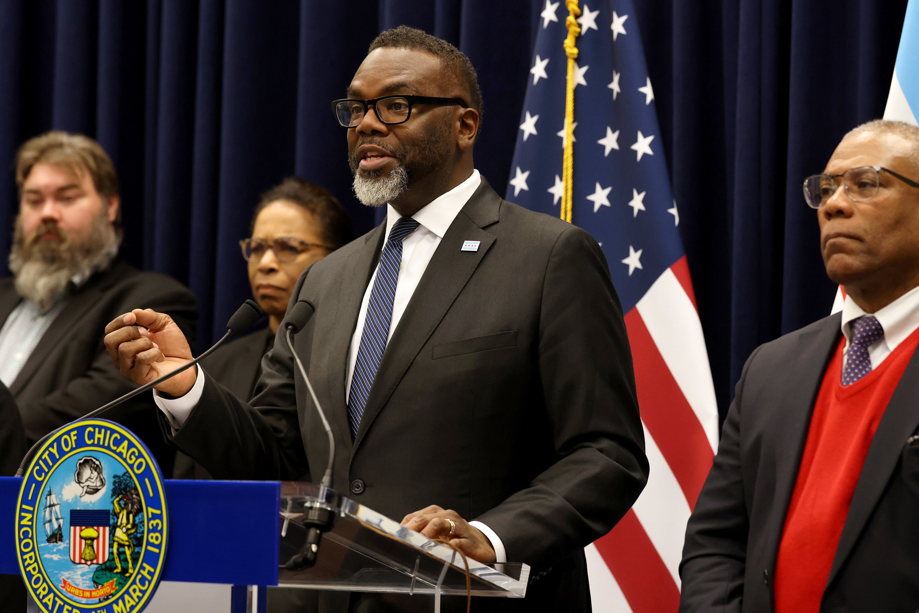 A man in a suit speaks behind a podium and in front of a row of three people in suits and an American flag and a black background.