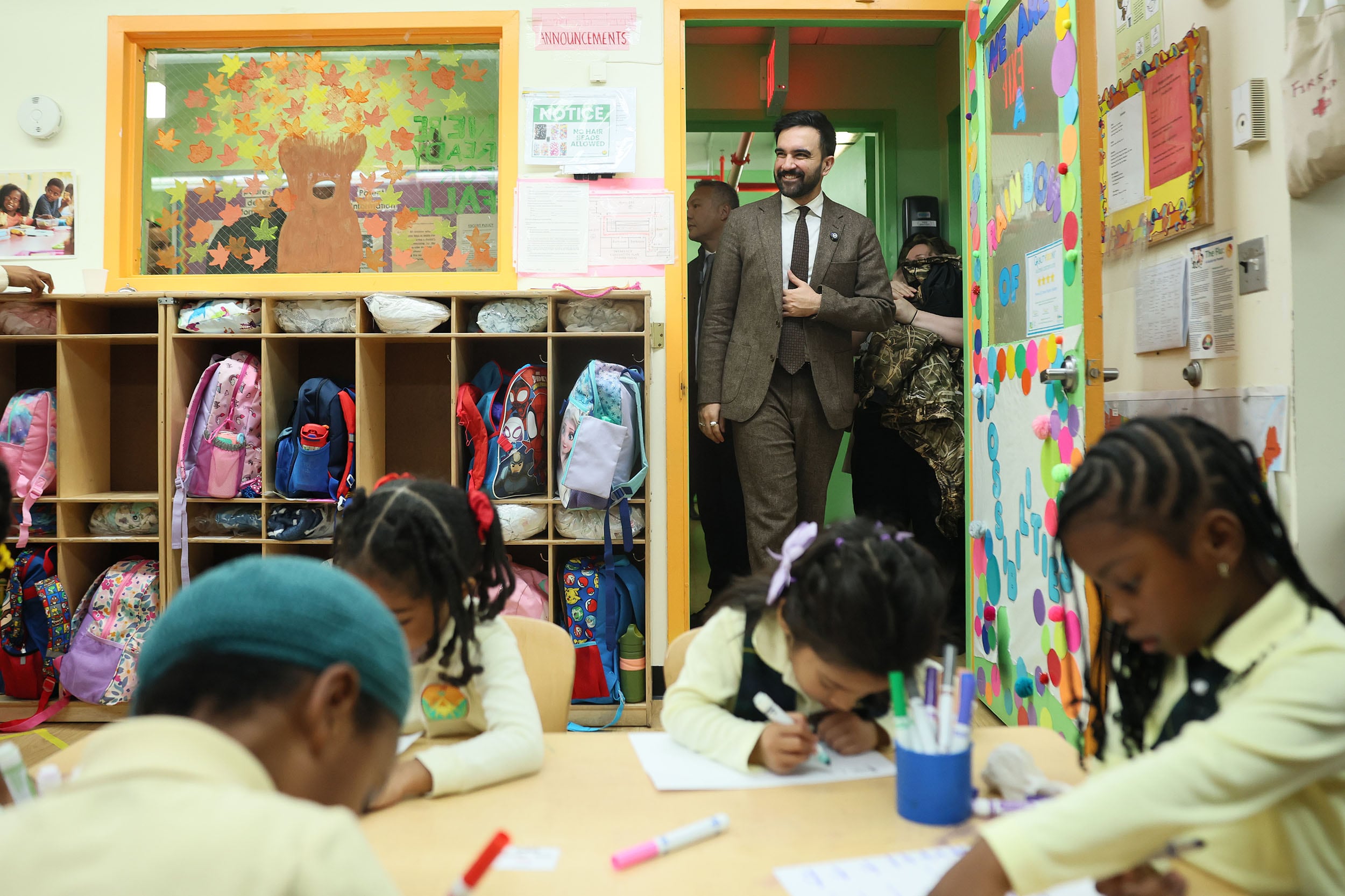 A photograph of a man in a suit walking through a door of a Pre-K classroom with four young students sitting a desk coloring.