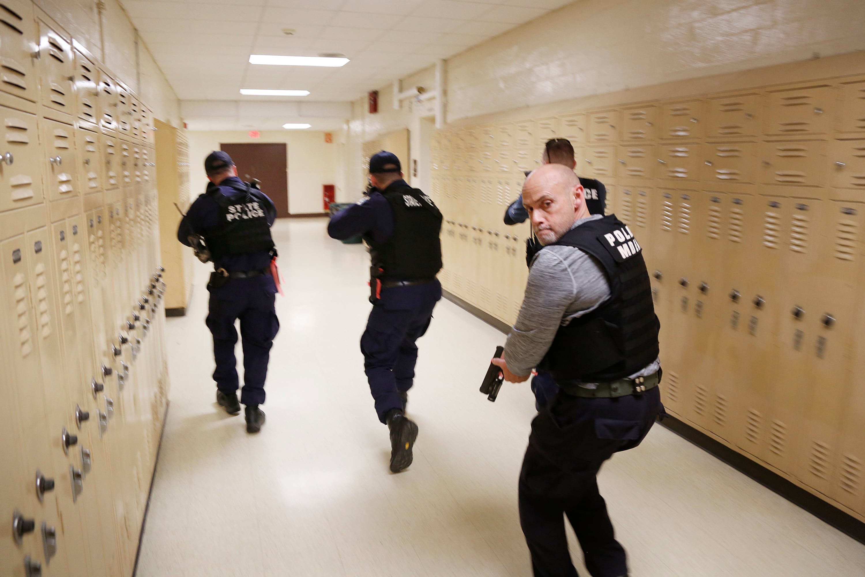 Four state police officers in bulletproof vests walk through a locker-lined school corridor with guns drawn during active-shooter training.