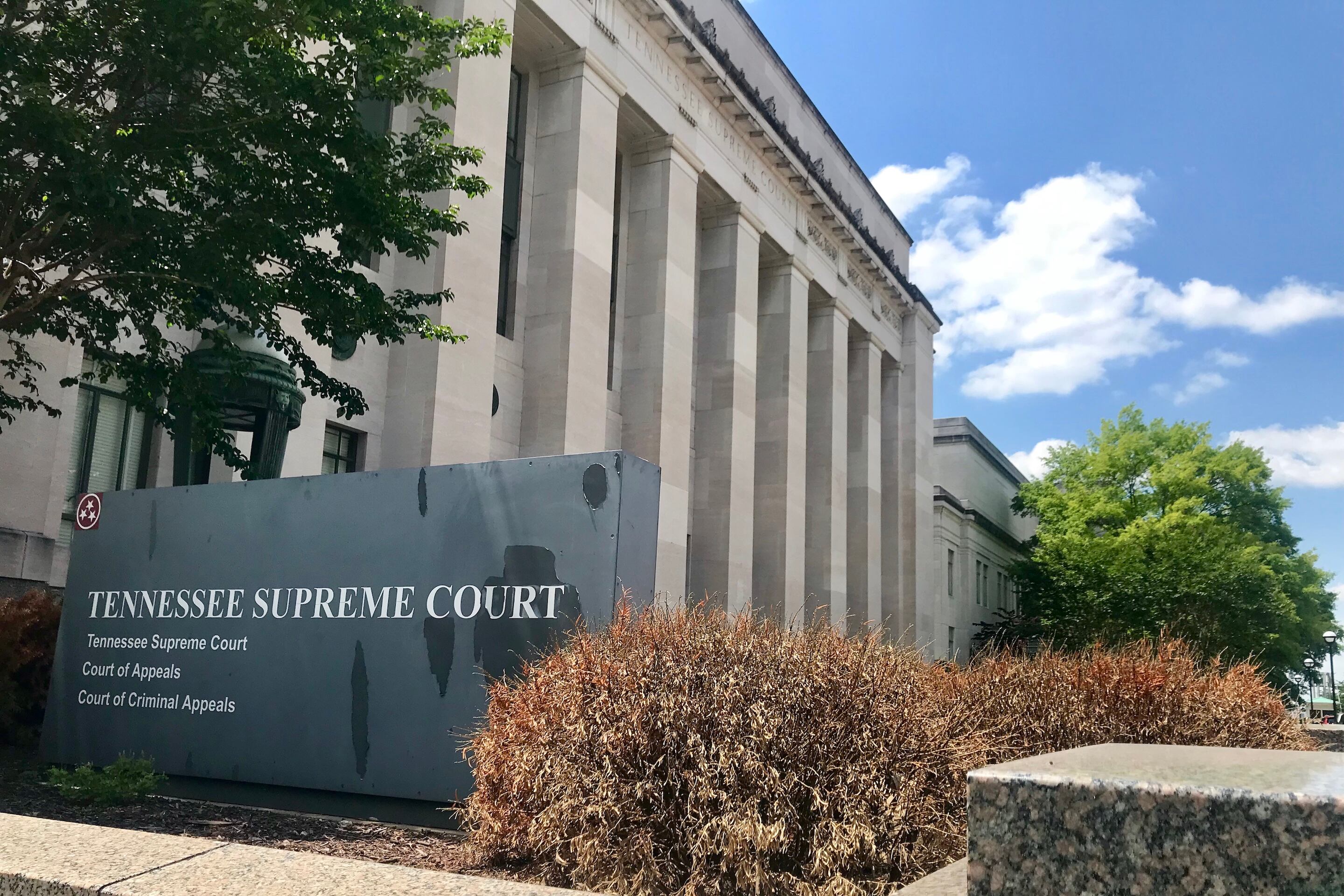 A large, multi-columned stone building set against a bright blue sky. A sign in front of the building identifies it as the Tennessee Supreme Court.