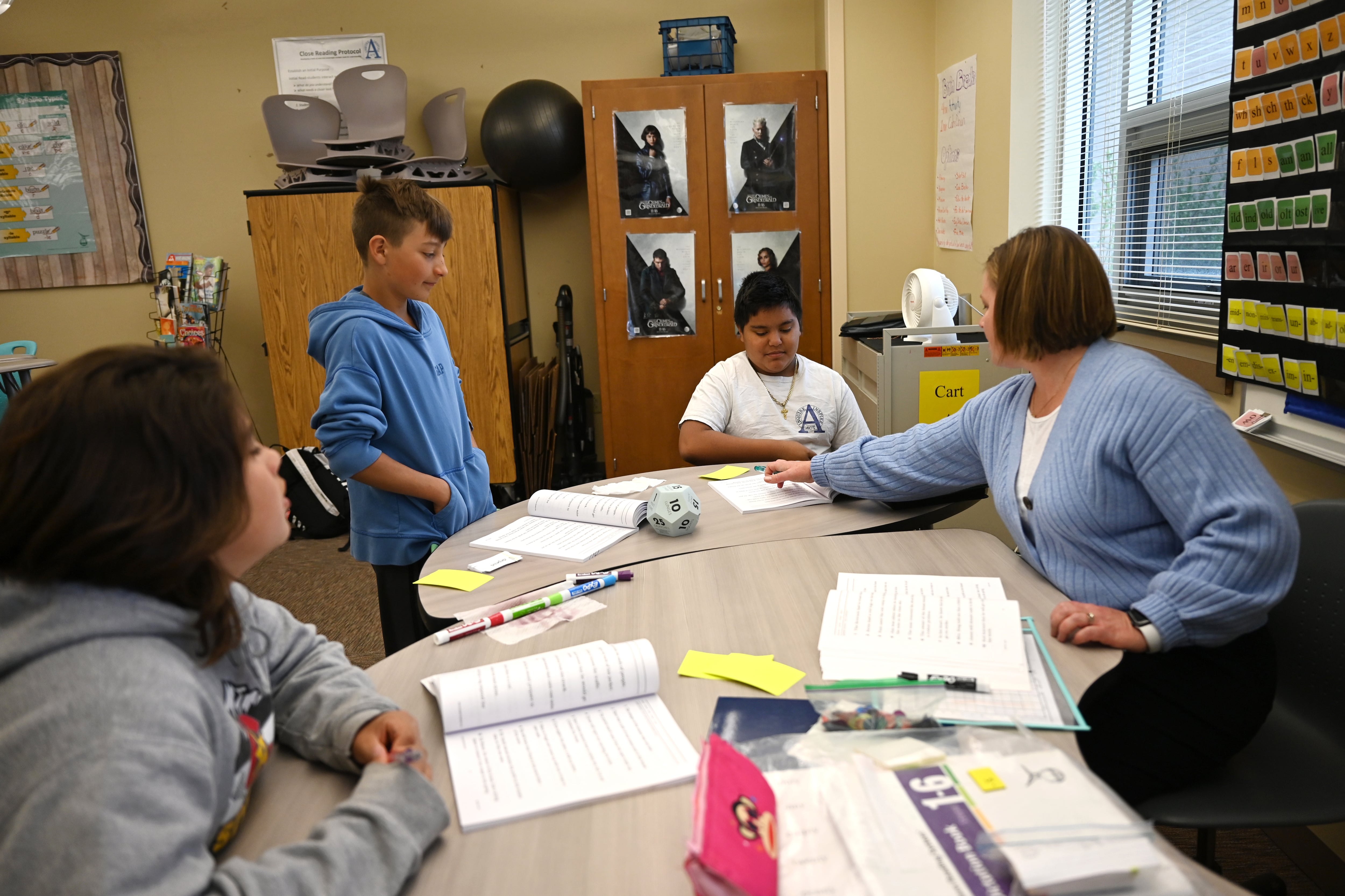 A teacher in a blue sweater works with three middle school boys on reading activities at a semi-circular table.