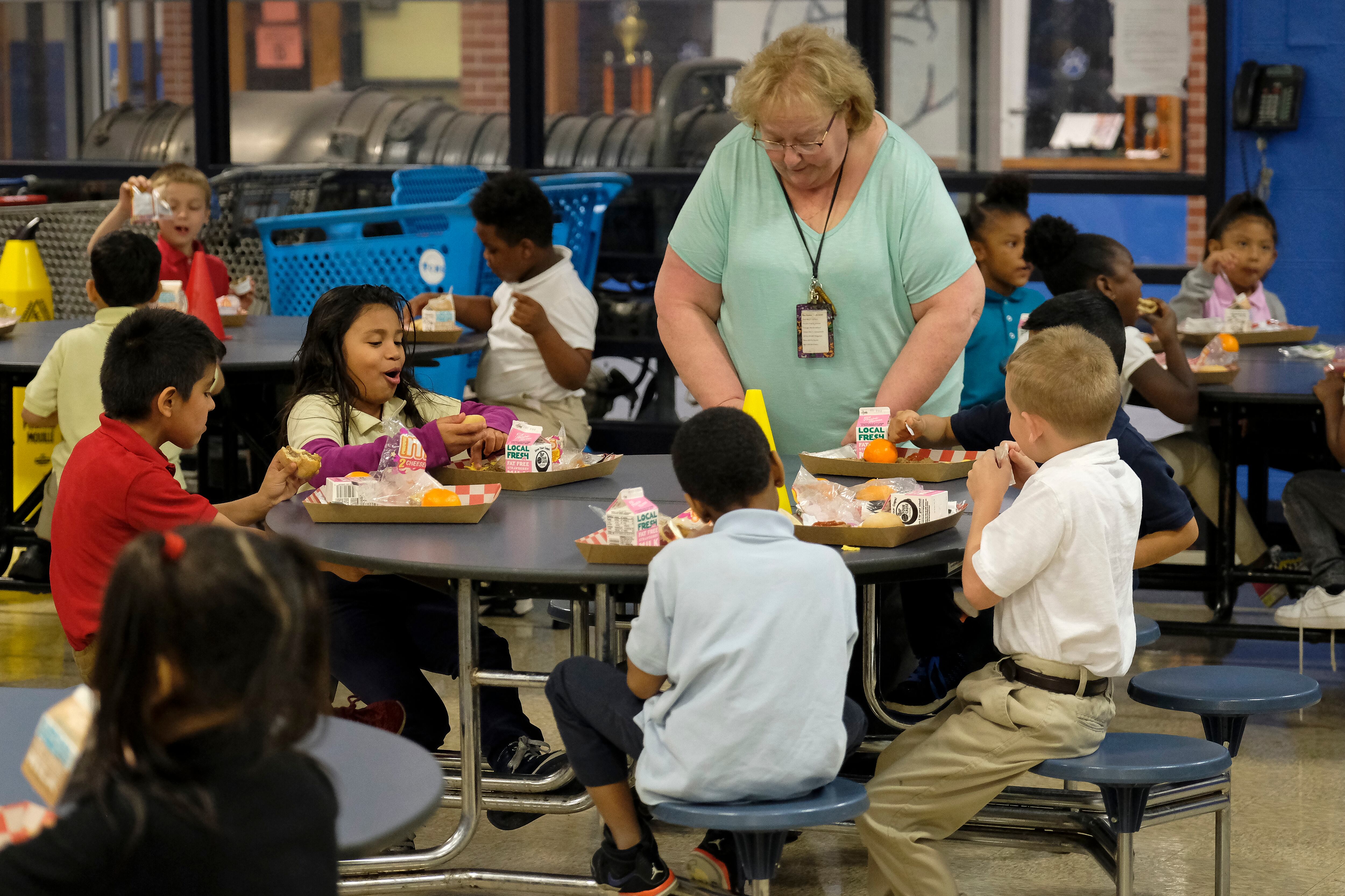 Students eat lunch as a older woman stands at their table in the cafeteria.