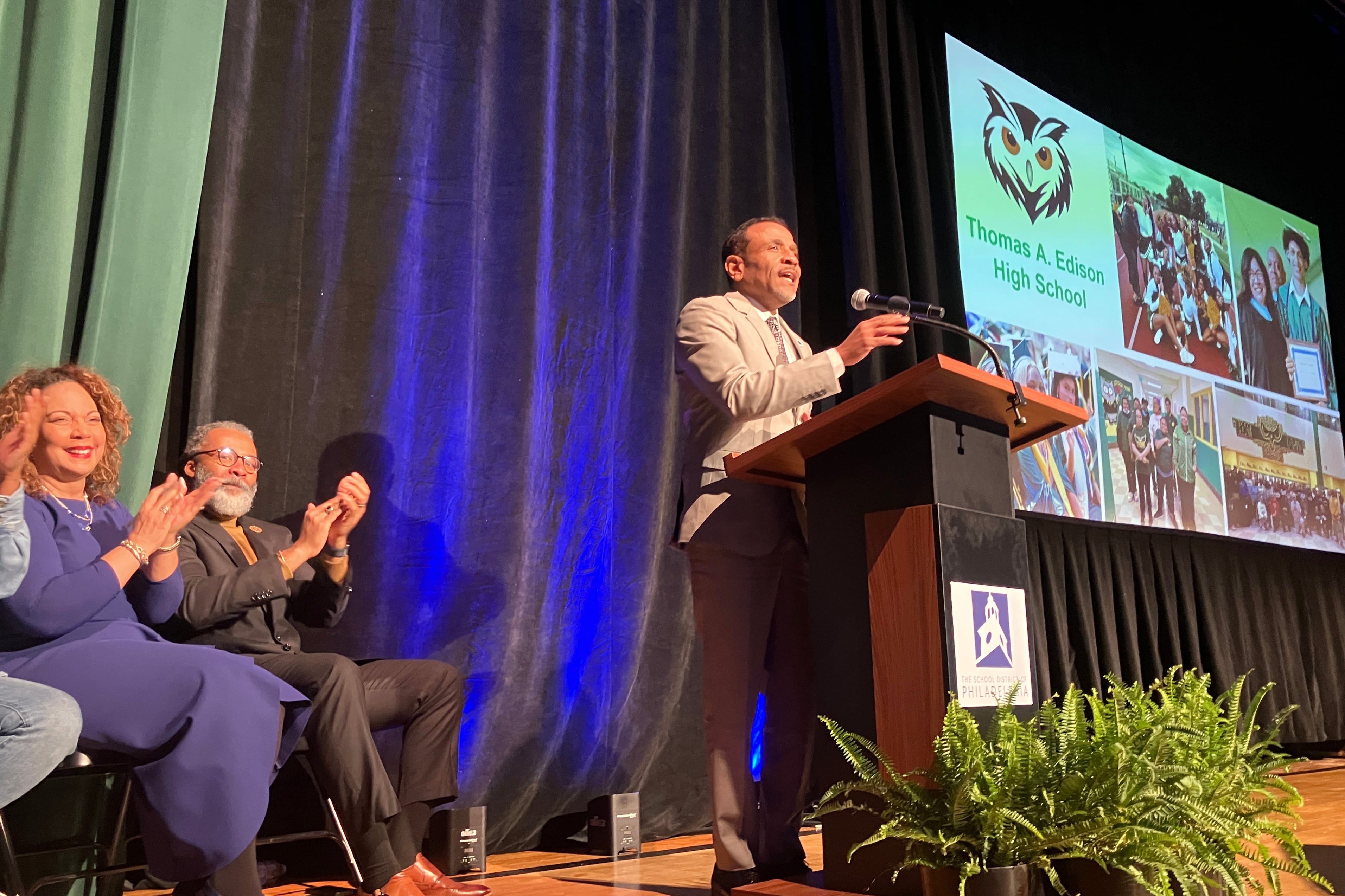 A photograph of a Black man in a suit speaking from behind a podium on a school auditorium stage.