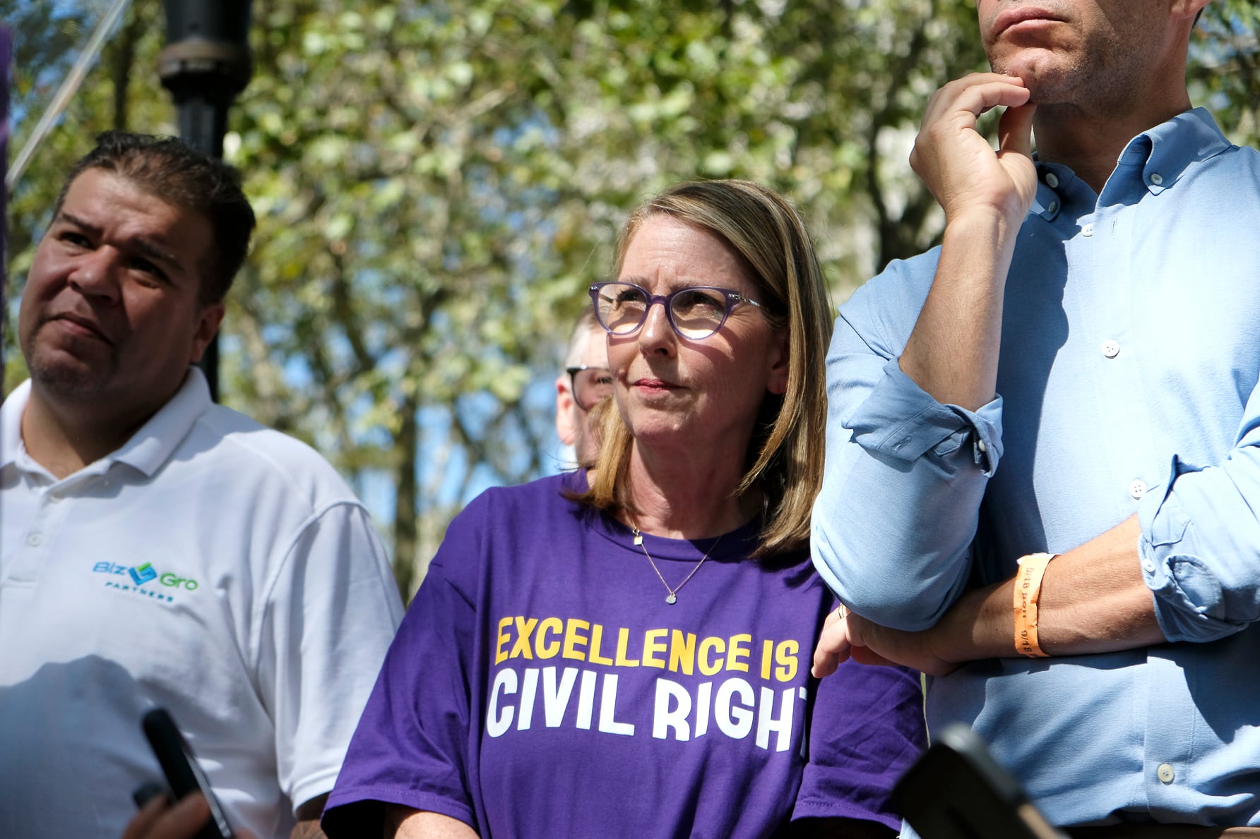 A woman in a purple t-shirt and glasses stands between two men in light shirts.